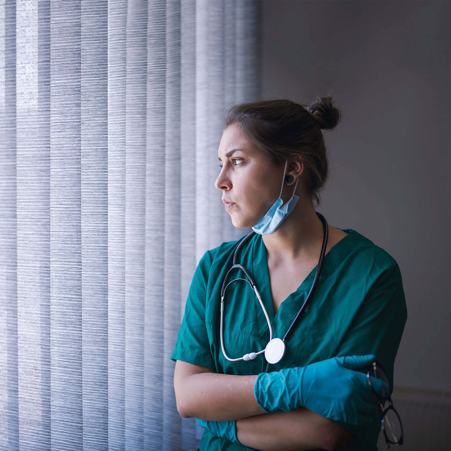 Nursing 桌布 壁紙 — 一位疲憊的護士穿著綠色手術衣和藍色手套，站在有垂直百葉窗的窗邊，頸上掛著聽診器，若有所思地望向窗外。