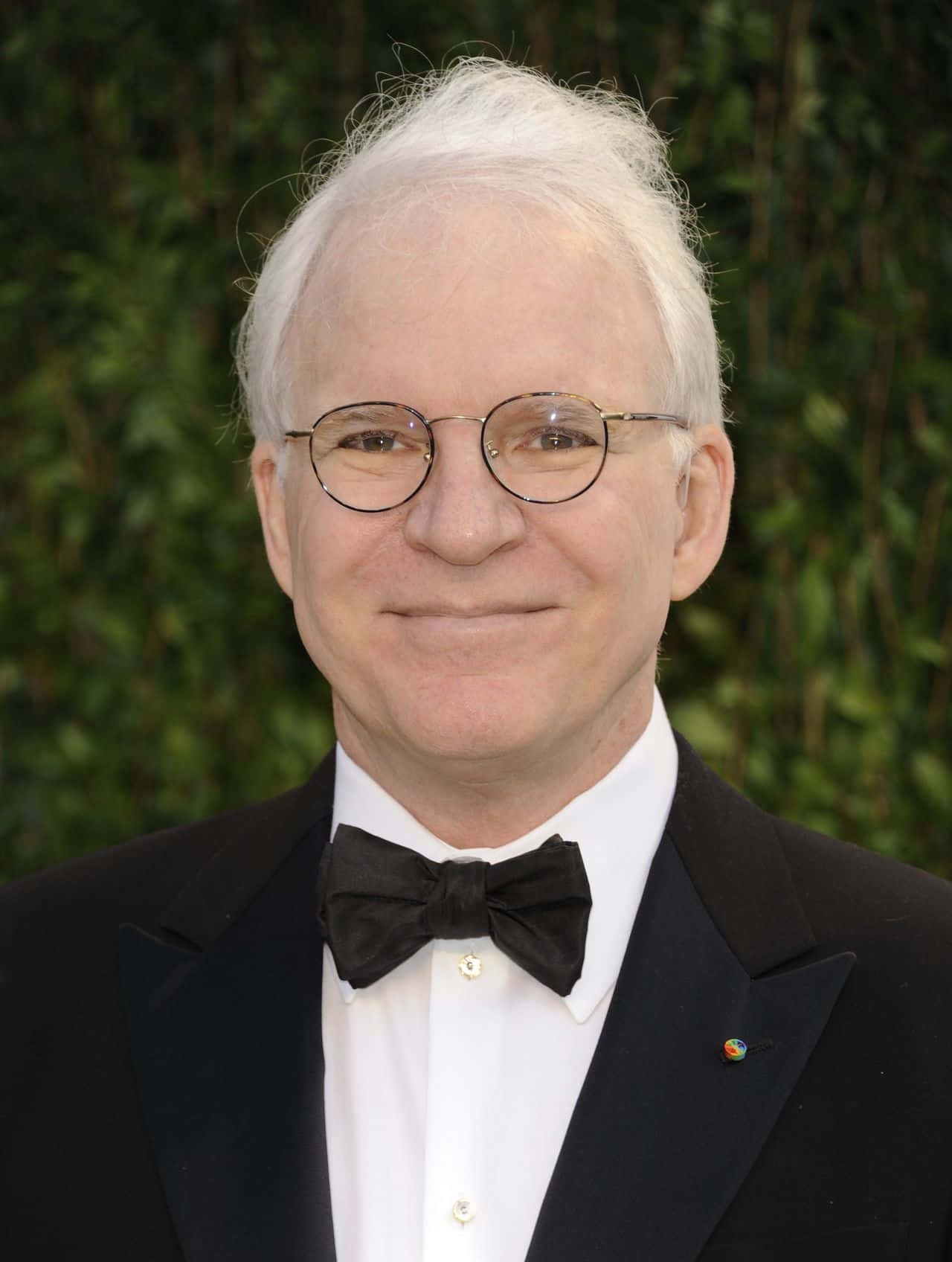 steve martin 桌布 壁紙 — 一位白髮、戴圓框眼鏡的男子，身穿黑色燕尾服配蝴蝶結，在綠色樹籬背景前微笑。