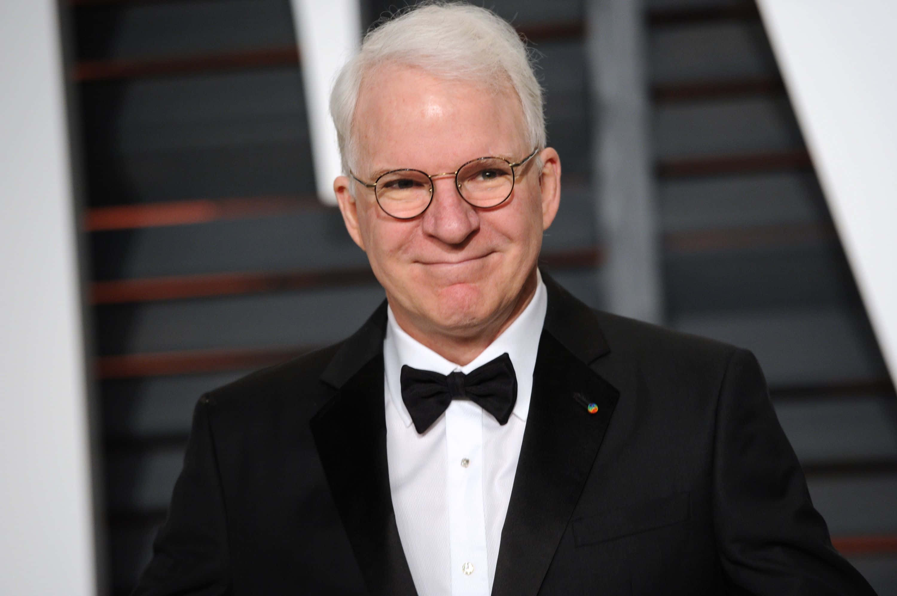 steve martin 桌布 壁紙 — 一位白髮戴眼鏡的微笑男士，身穿黑色燕尾服和領結，背景為幾何條紋。