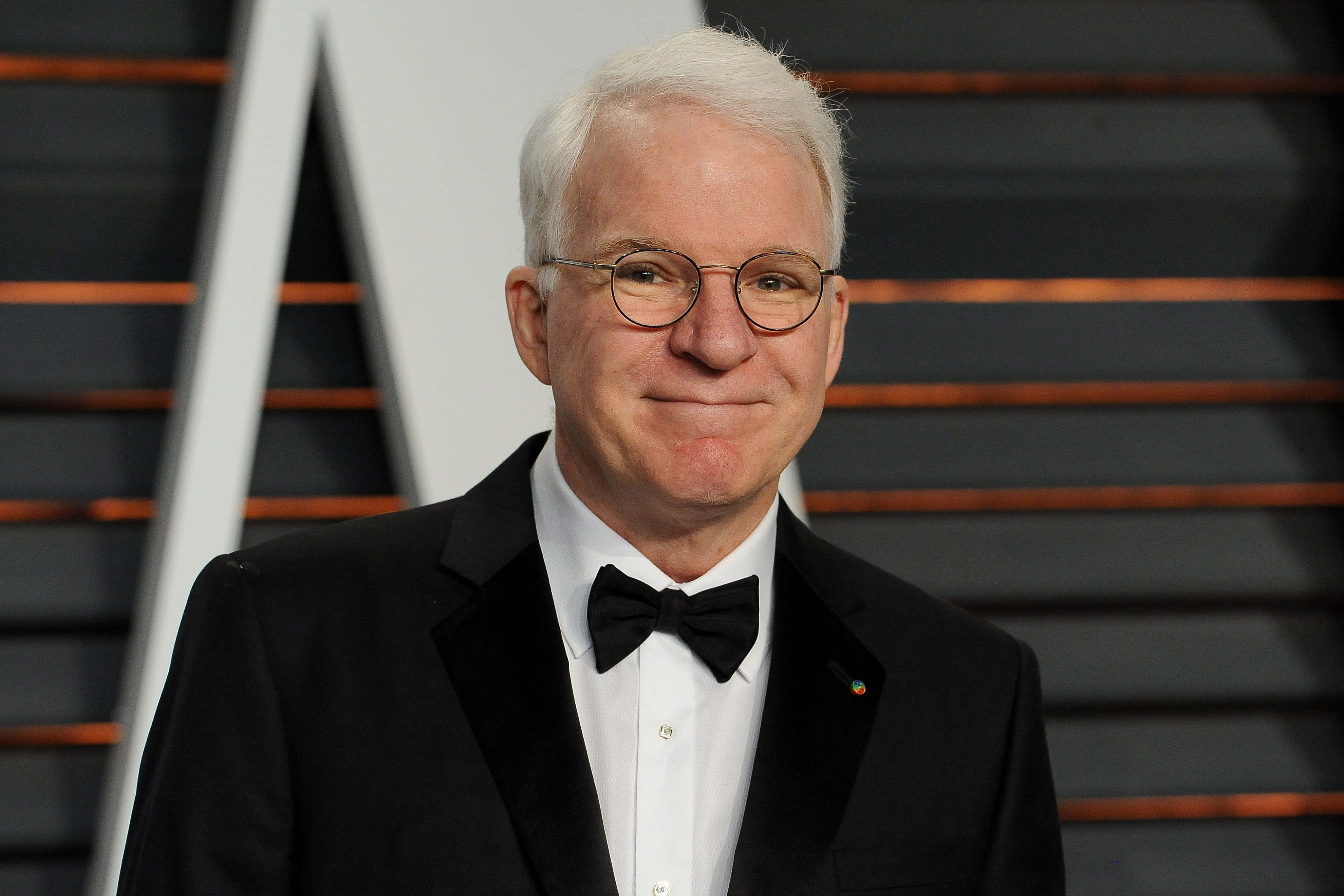 steve martin 桌布 壁紙 — 一位白髮戴眼鏡的男子，身穿黑色燕尾服，在條紋背景前微笑。