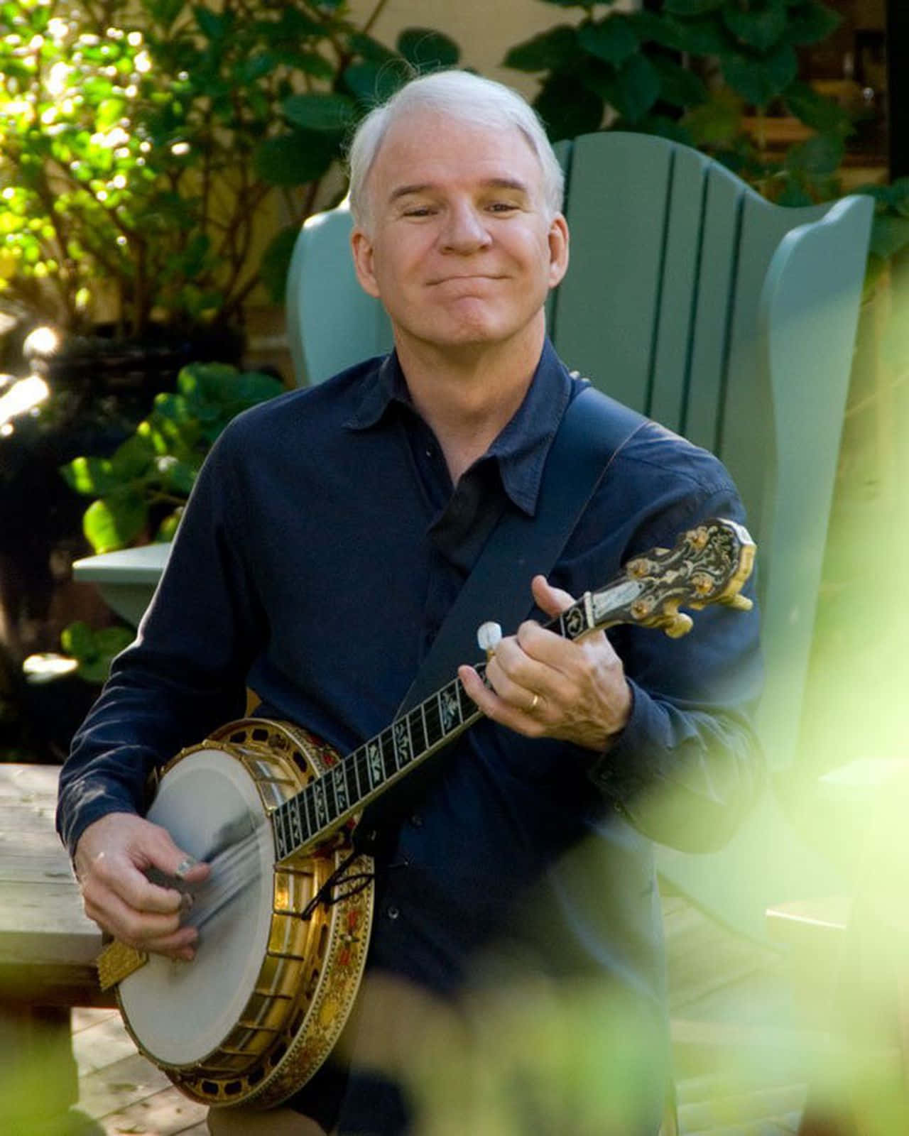 steve martin 桌布 壁紙 — 一位白髮男士在陽光明媚的花園椅上微笑，手持裝飾性班卓琴。