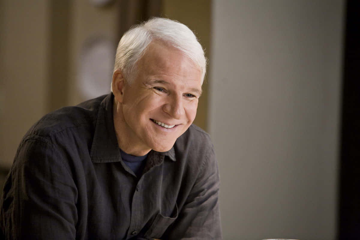 steve martin 桌布 壁紙 — 一位白髮的開朗長者，在室內穿著深色鈕扣襯衫，露出溫暖的笑容。