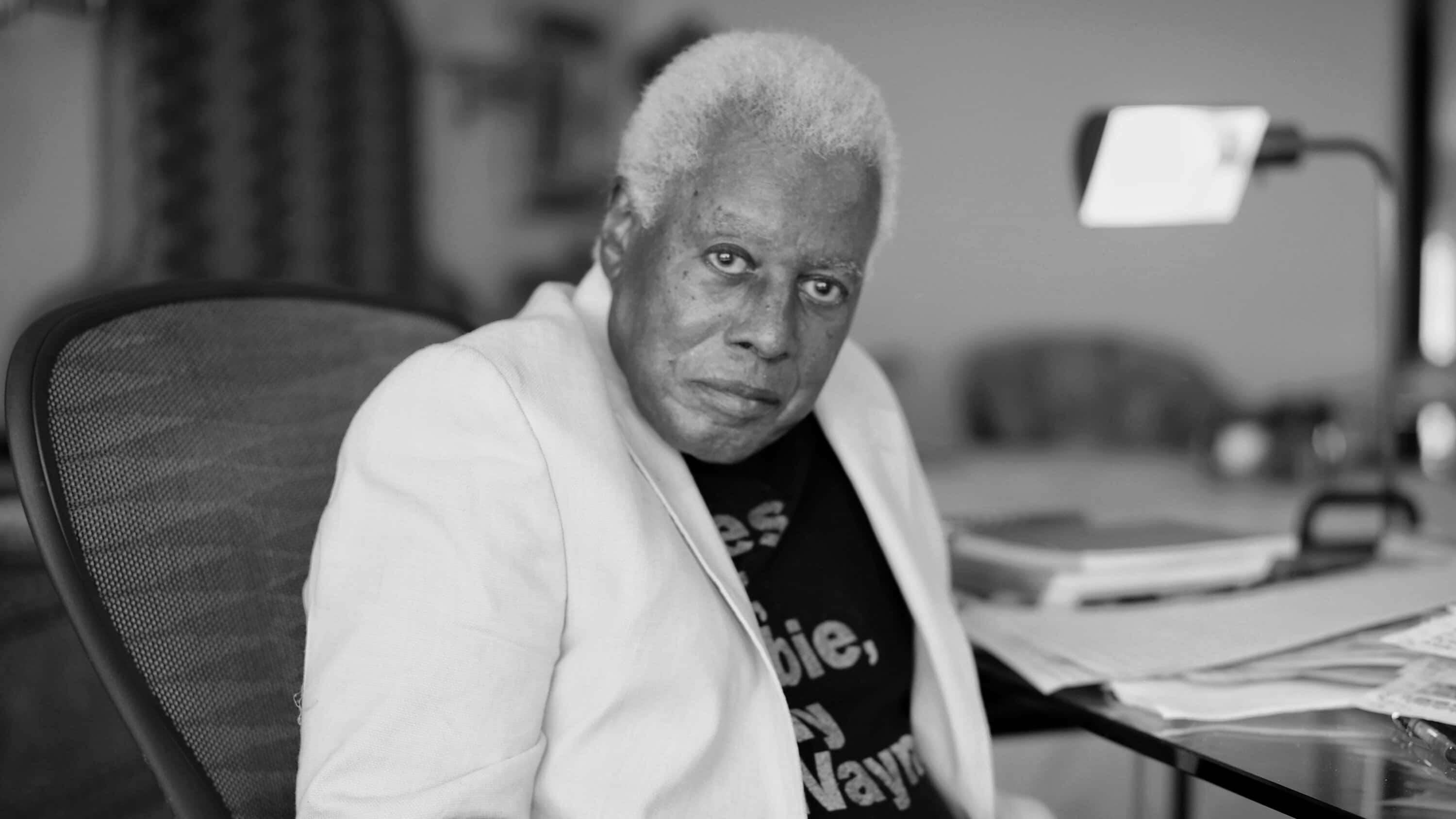 Wayne Shorter 桌布 壁紙 — 一位白髮老人在燈光昏暗的辦公室裡坐在雜亂的辦公桌前的黑白肖像。