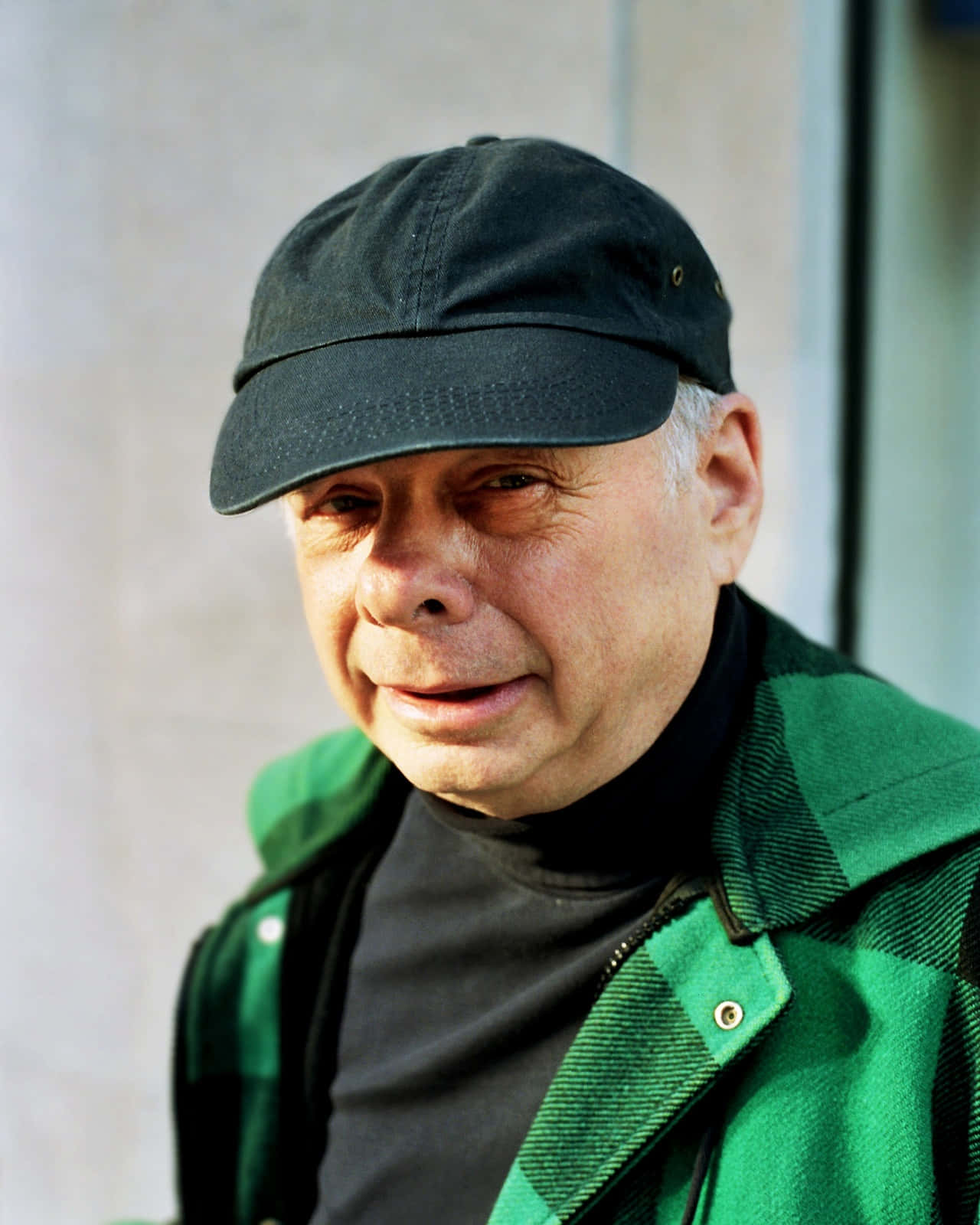 Wallace Shawn 桌布 壁紙 — 一位白髮老翁戴著黑色鴨舌帽，身穿綠色格仔外套，靠著中性色調的牆壁。