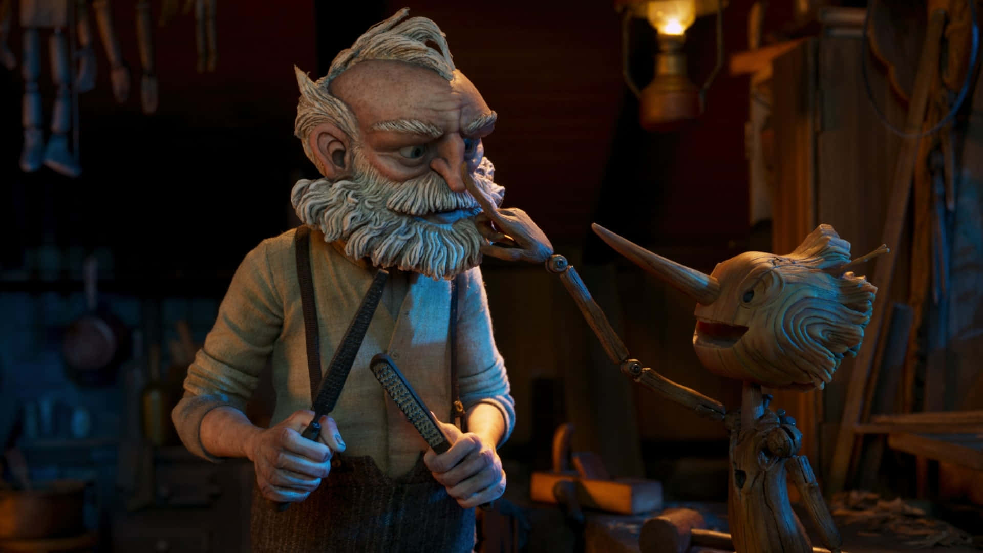 Guillermo Del Toro's Pinocchio 桌布 壁紙 — 一位白鬍鬚灰白頭髮的木雕師傅在光線昏暗、工具滿佈的工作室裡小心翼翼地雕刻著皮諾丘的鼻子。