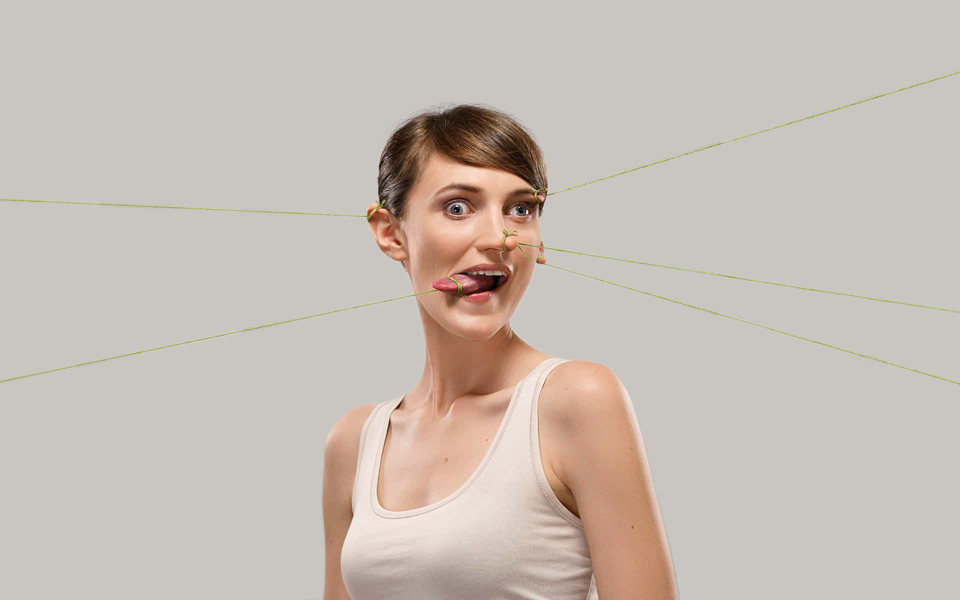 Funny Faces 桌布 壁紙 — 一位眼睛睜大、嘴巴張開的女性，臉上繫著細細的綠色繩子，在純灰色背景上拉緊。