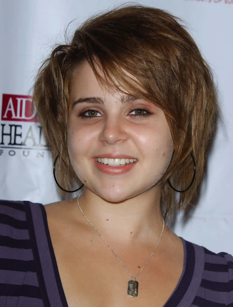Mae Whitman 桌布 壁紙 — 一位短棕髮、戴耳環的年輕女性，穿著條紋上衣，對著鏡頭溫暖微笑。