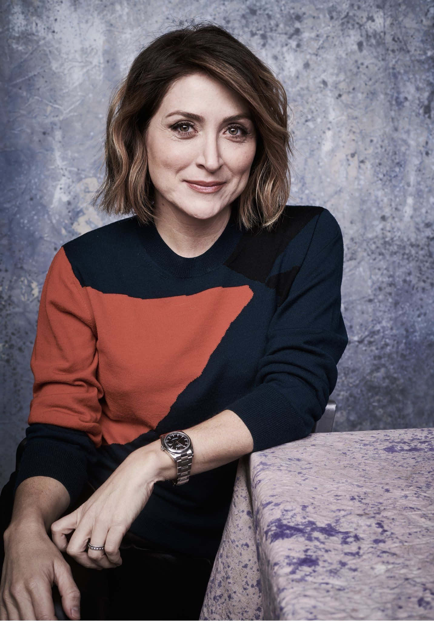 Sasha Alexander 桌布 壁紙 — 一位短棕髮女性，穿著拼色毛衣，在紋理灰色背景前溫柔微笑。