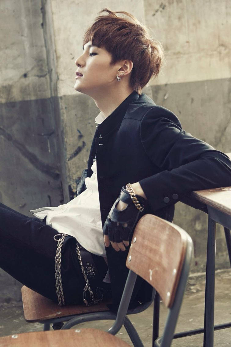 BTS SUGA《Skool Luv Affair》 壁紙 — 一位短棕髮的年輕男子隨意地坐在木椅上，背景是風化的混凝土牆，身穿黑色外套和帶鏈條的白襯衫。