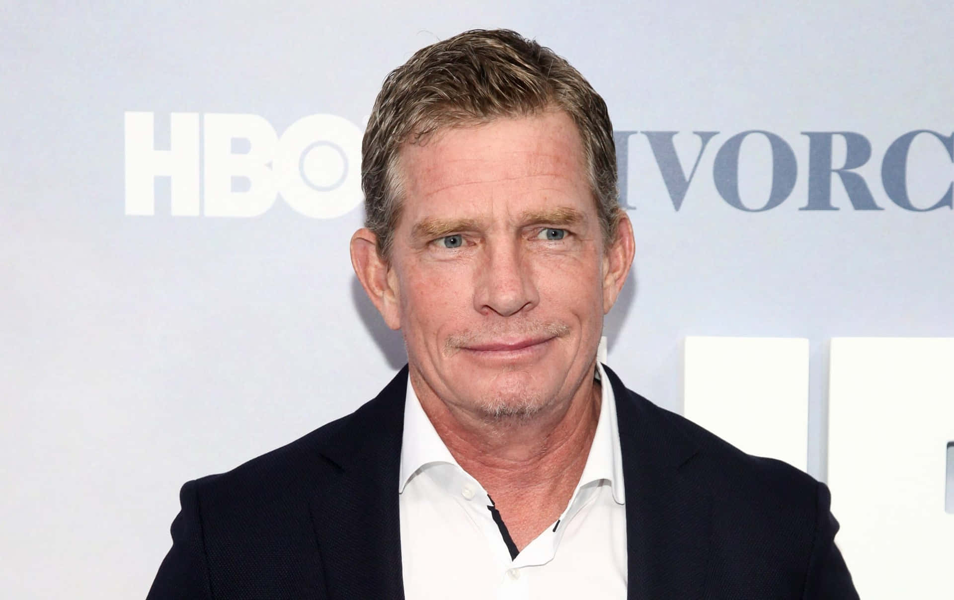 Thomas Haden Church 桌布 壁紙 — 一位短棕髮藍眼的男士，身穿深色西裝和白襯衫，背景為HBO。