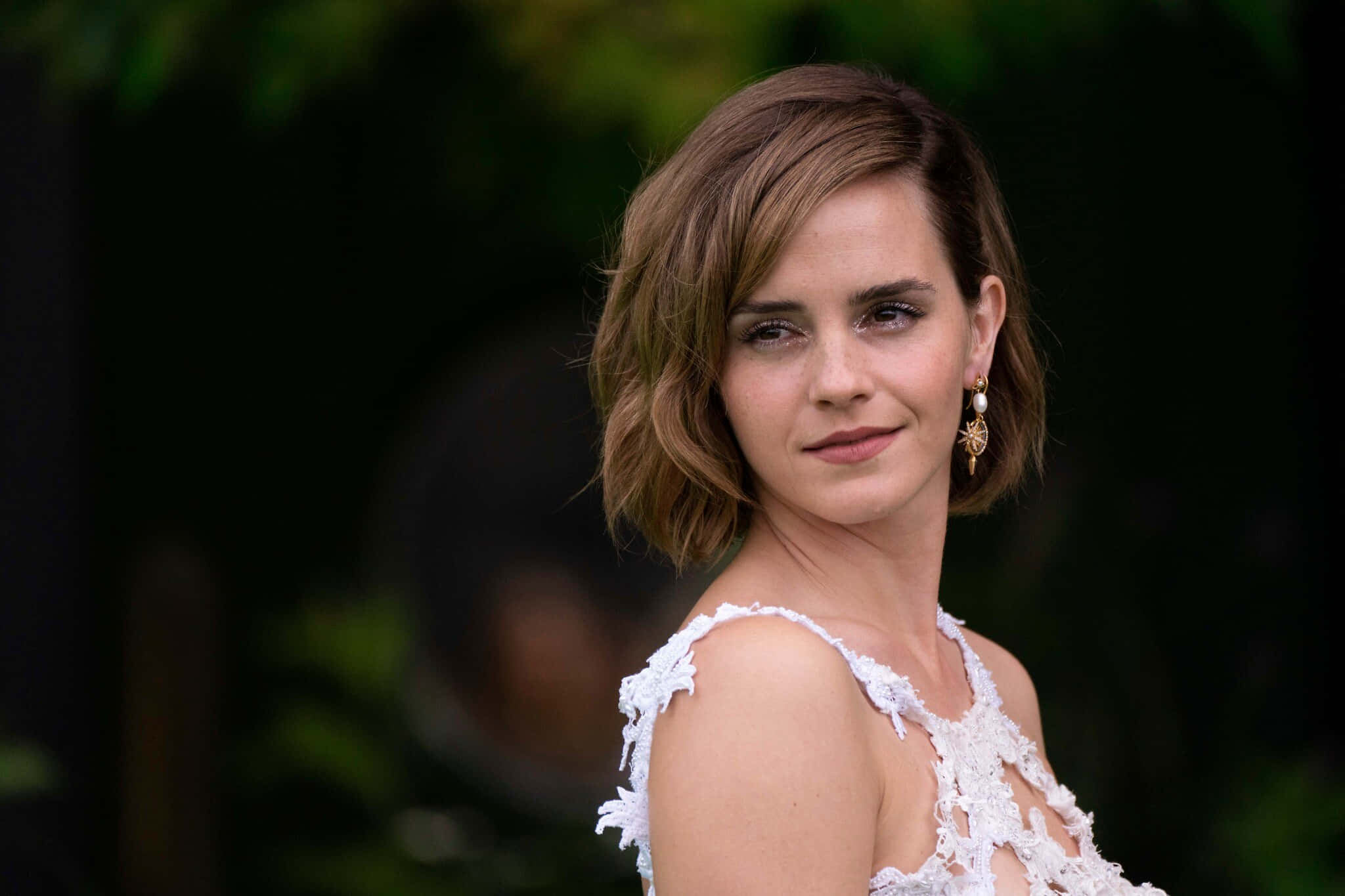 Emma Watson 桌布 壁紙 — 一位短波浪棕色頭髮的女性，在模糊的綠色背景前穿著白色蕾絲上衣和金色耳環。