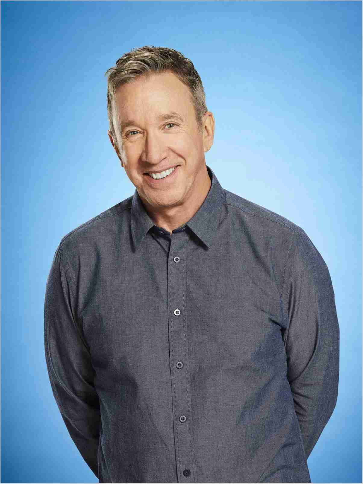 Tim Allen 桌布 壁紙 — 一位短灰髮的開朗男士穿著深灰色鈕扣襯衫，在純藍色攝影棚背景前溫暖地微笑。