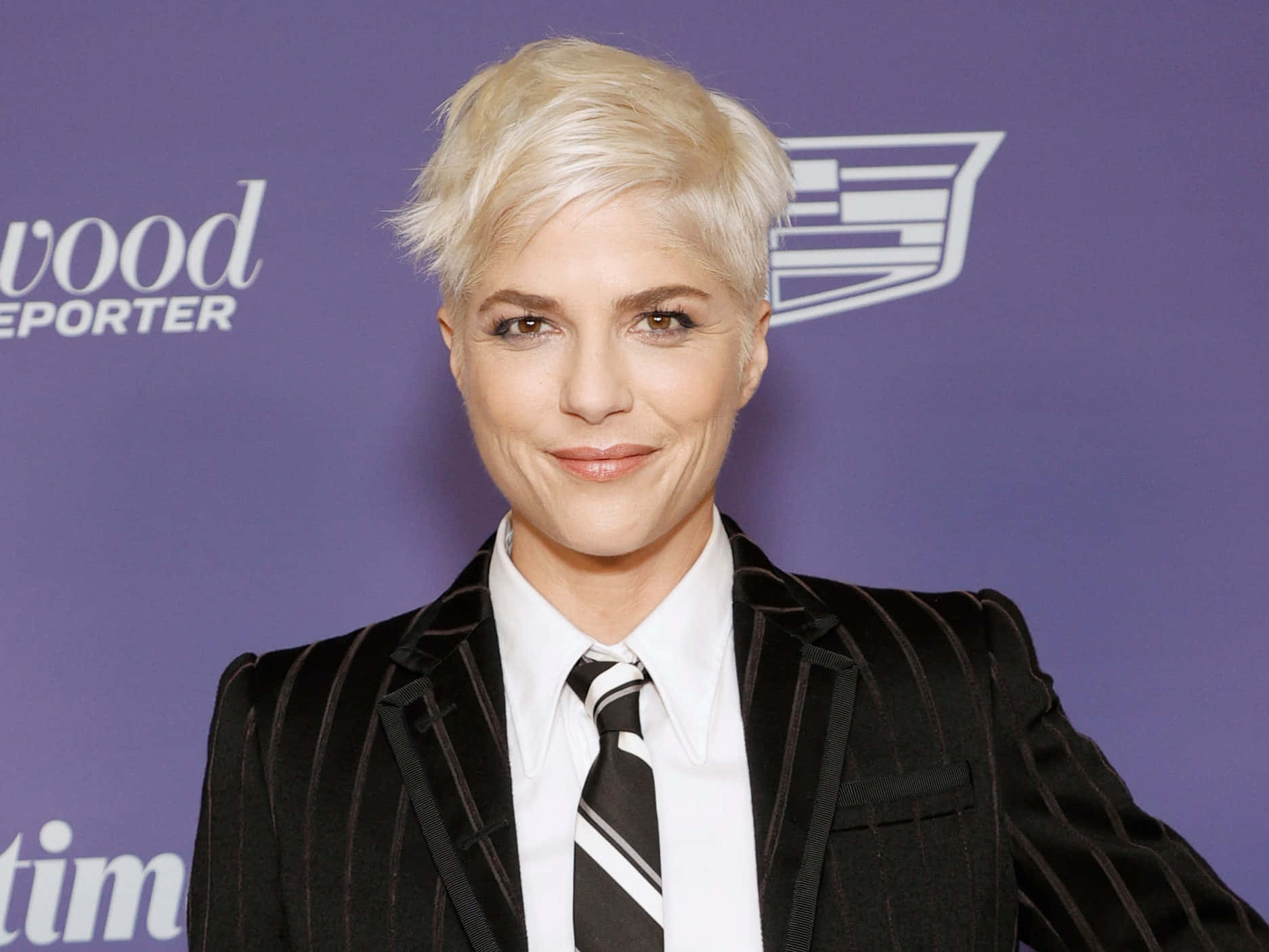 Selma Blair 桌布 壁紙 — 一位短白金髮女性穿著黑色細條紋西裝，在紫色活動背景前自信微笑。