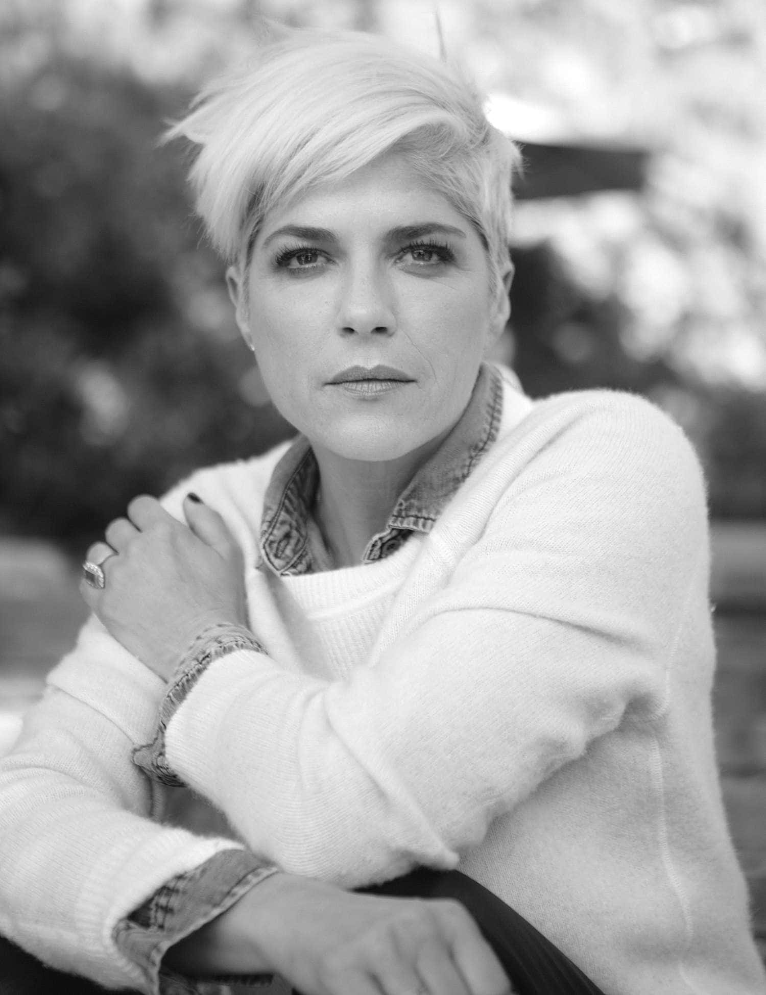 Selma Blair 桌布 壁紙 — 一位短白金髮的女性，身穿柔軟毛衣搭配牛仔襯衫，以黑白風格沉思地坐著。