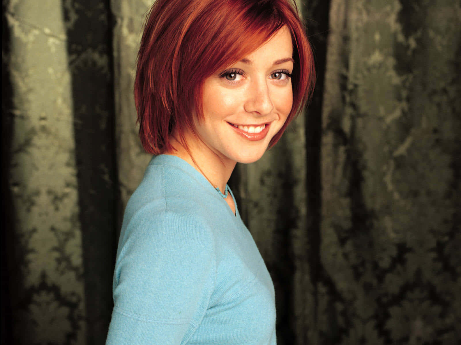 Alyson Hannigan 桌布 壁紙 — 一位短紅髮女性穿著淺藍色毛衣，在深色圖案窗簾背景下溫暖微笑。