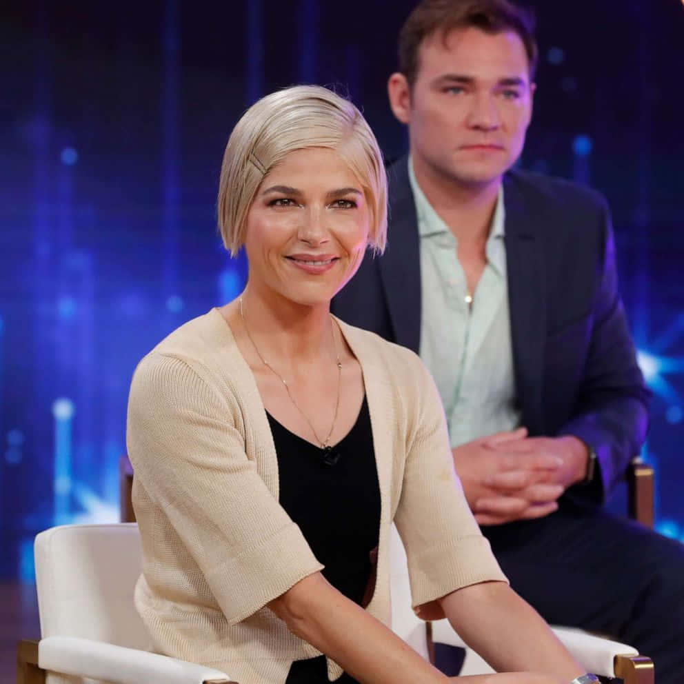 Selma Blair 桌布 壁紙 — 一位短金髮女性坐在白色椅子上，在背景有男士的攝影棚訪談中溫暖地微笑。