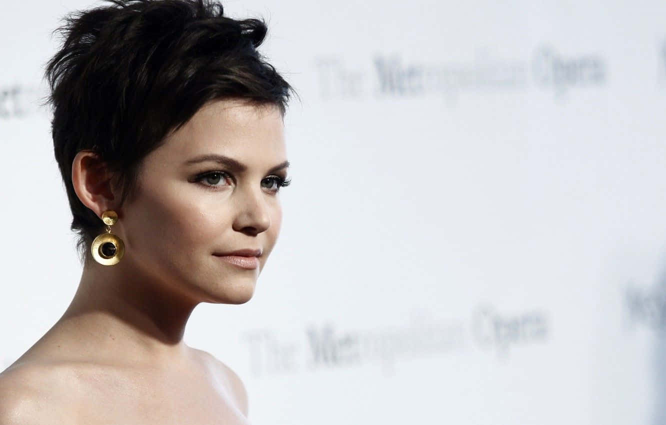 ginnifer goodwin 桌布 壁紙 — 一位短黑髮、戴金色圓環耳環的女性，在模糊的「大都會歌劇院」背景下若有所思地凝視。