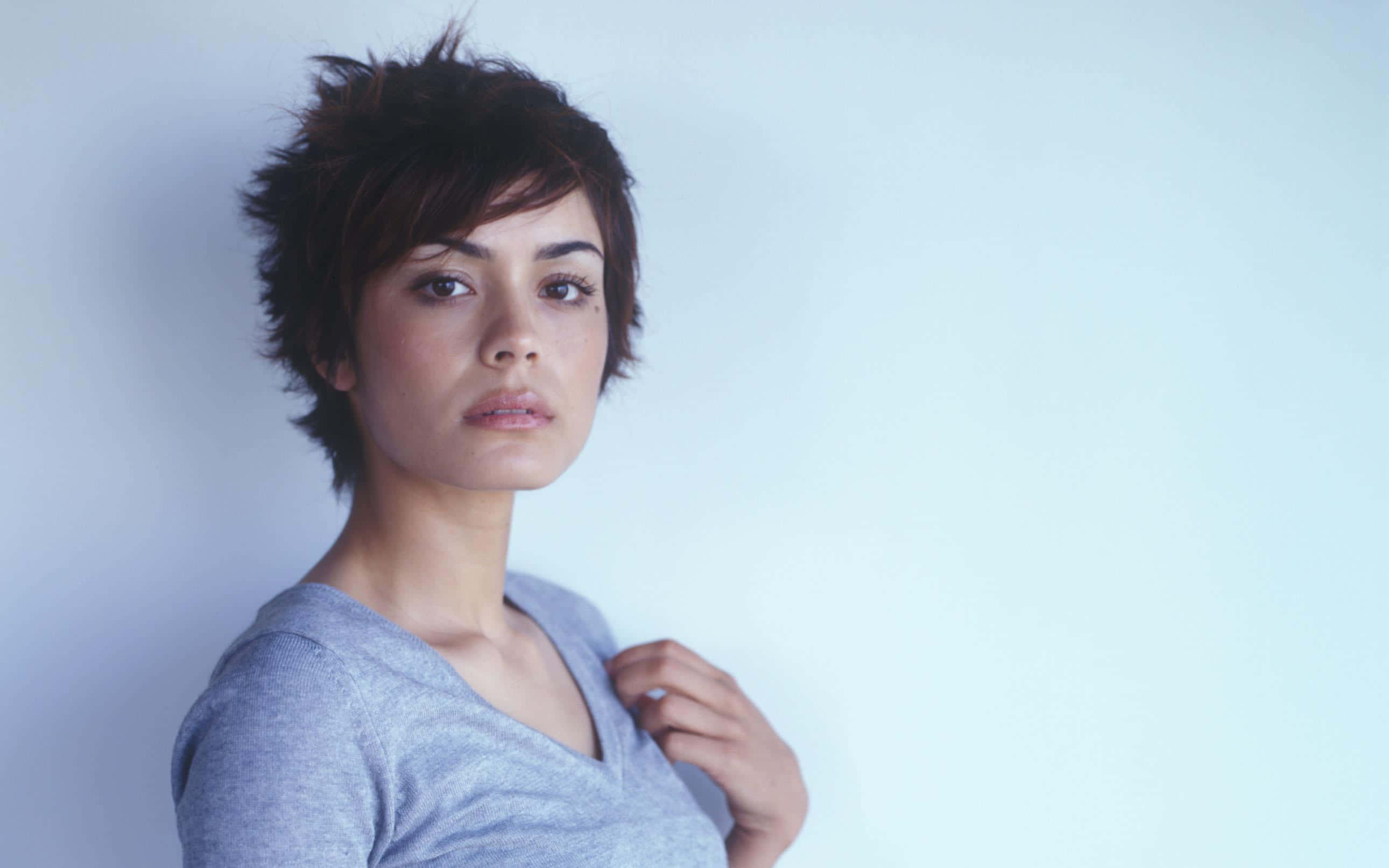 Shannyn Sossamon 桌布 壁紙 — 一位短黑髮、身穿灰色襯衫的女性，在柔和的藍色背景下若有所思地凝視著。