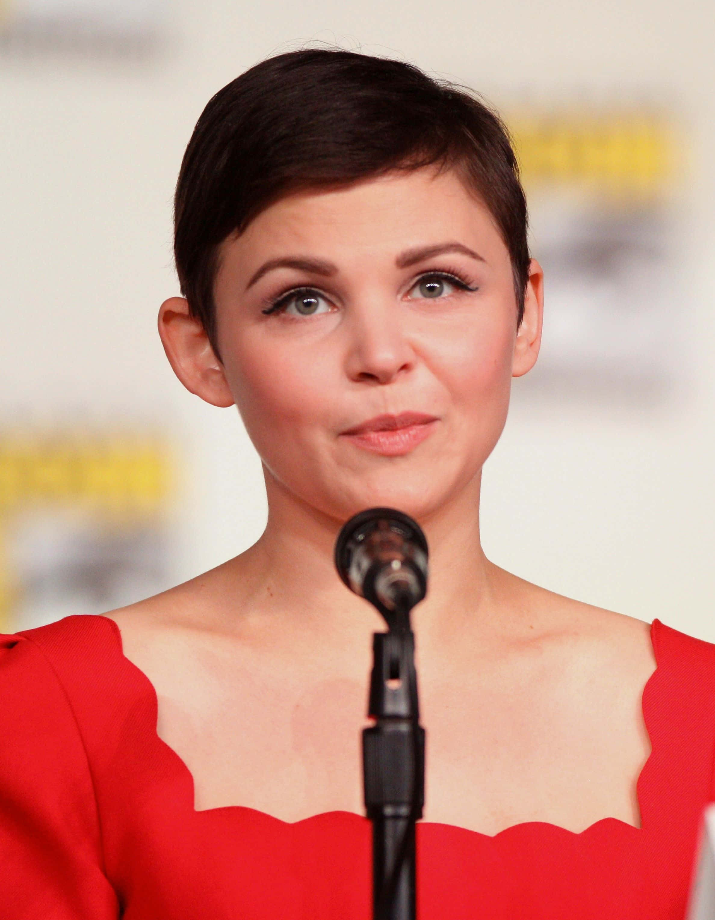 ginnifer goodwin 桌布 壁紙 — 一位短黑髮、身穿紅色連衣裙的女性在座談會活動中對著麥克風發言。