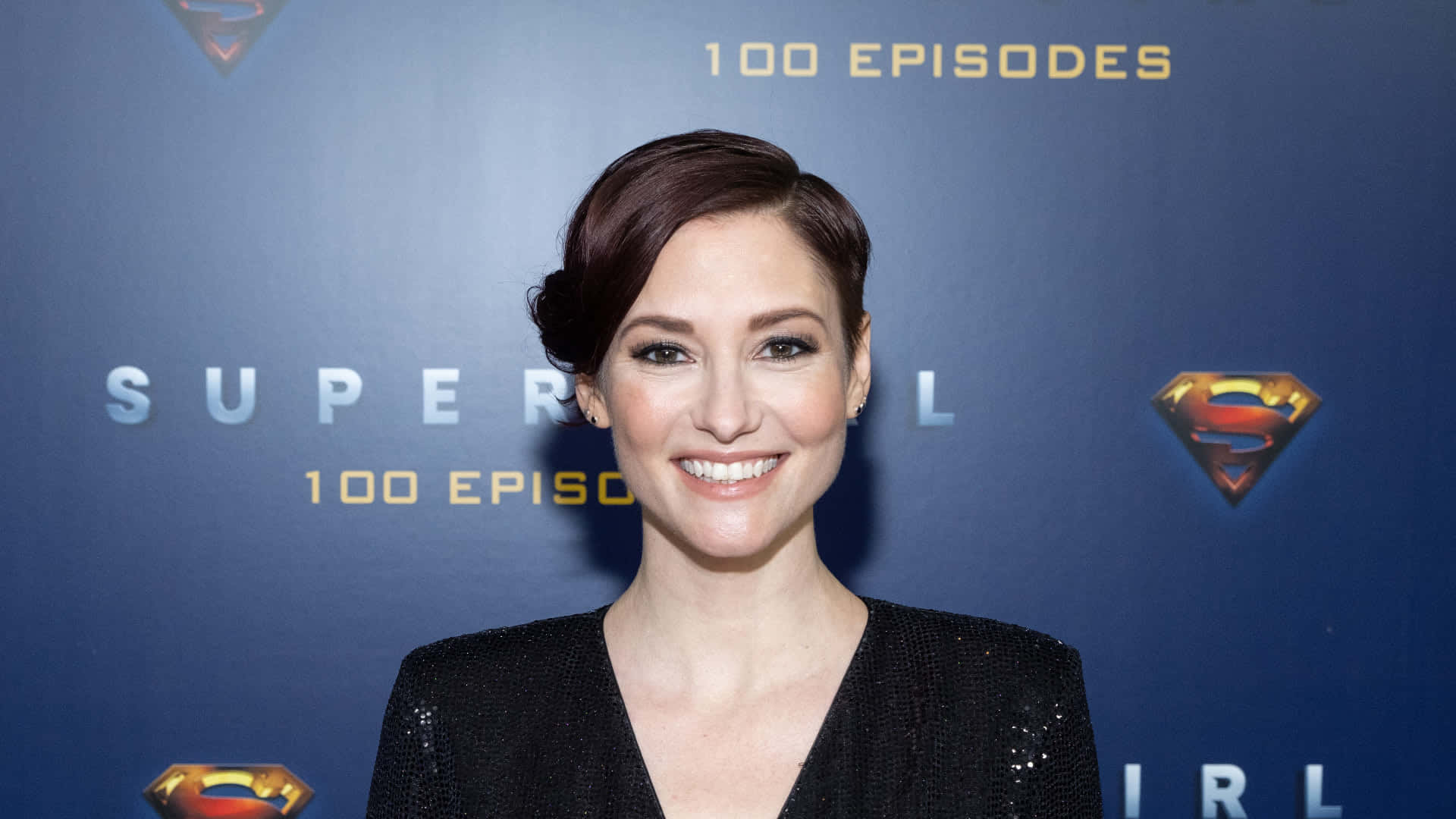 Chyler Leigh 桌布 壁紙 — 一位短黑髮、身穿黑色閃亮上衣的微笑女士站在藍色「Supergirl 100 Episodes」活動背景前。