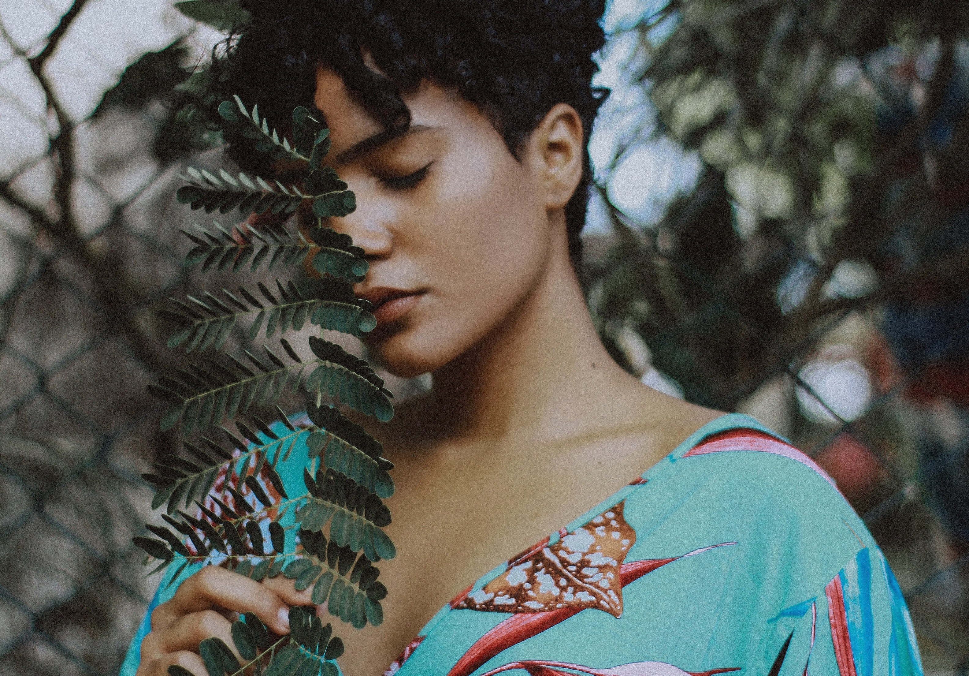 Beautiful Fashion 桌布 壁紙 — 一位短黑髮女性在模糊的鐵絲網背景前，輕輕將一片蕨葉靠近臉龐。