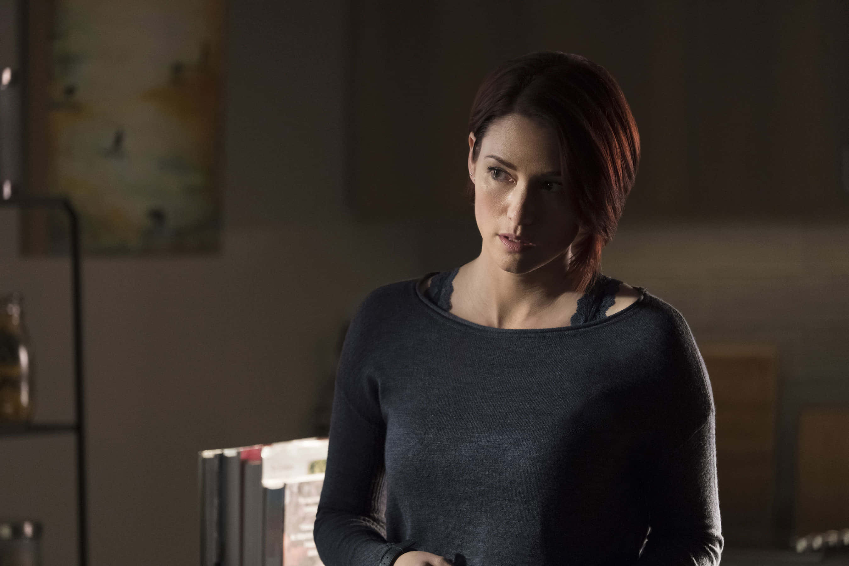 Chyler Leigh 桌布 壁紙 — 一位短黑髮女性站在燈光昏暗的房間裡，穿著帶有蕾絲邊的深色毛衣，在柔和的光線下。
