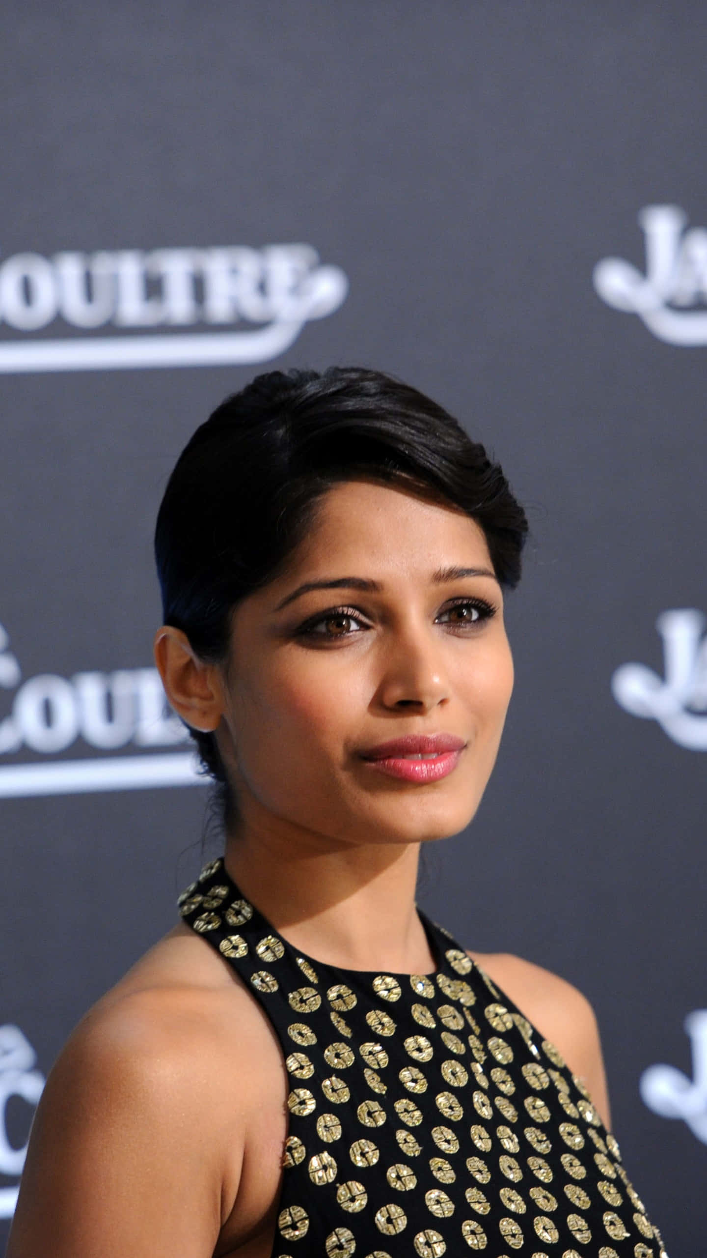 Freida Pinto 桌布 壁紙 — 一位短黑髮的優雅女士，身穿帶有金色圓形圖案的黑色連衣裙，背景為品牌背景。