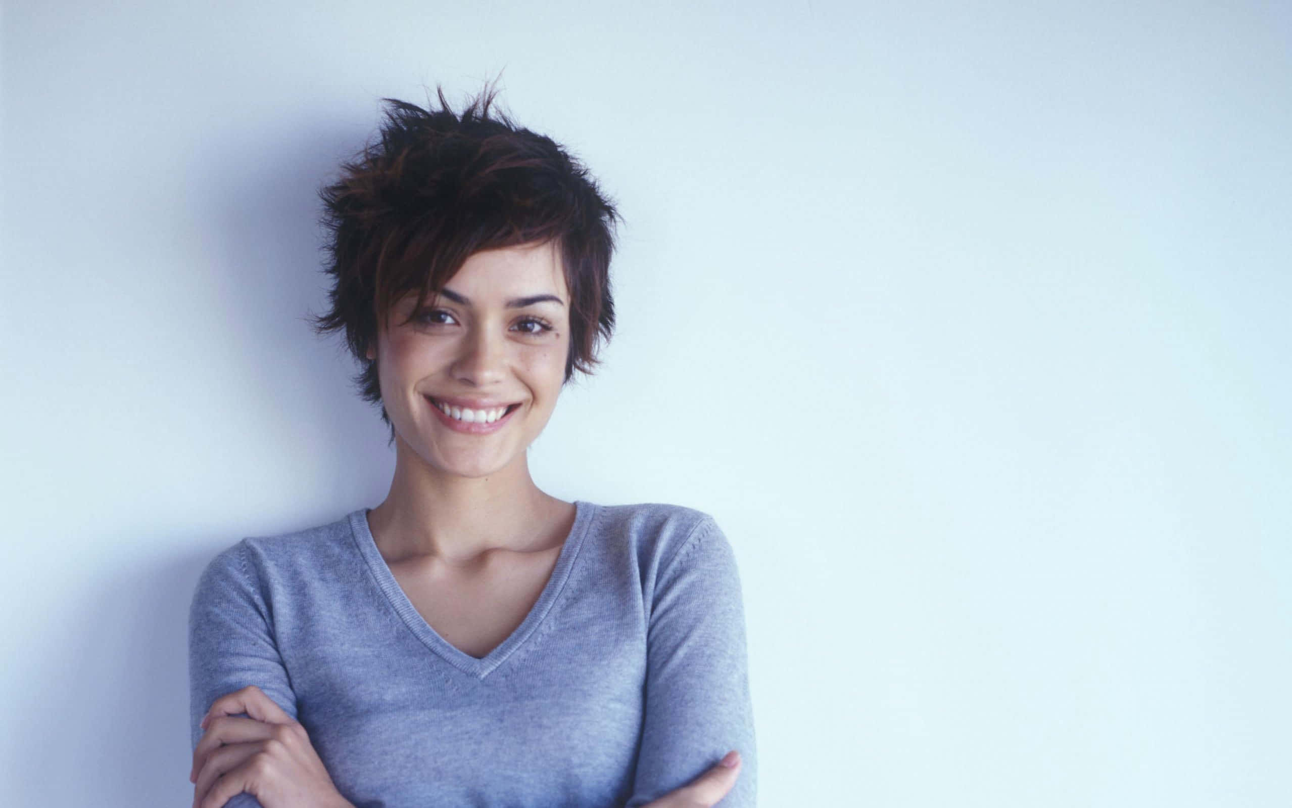 Shannyn Sossamon 桌布 壁紙 — 一位短黑髮的開朗女性靠著純淺藍色牆壁，露出溫暖的笑容。