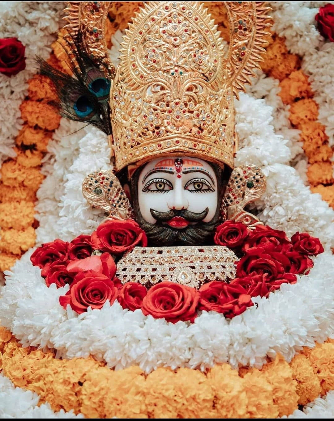 India 桌布 壁紙 — 一位神聖的神祇，頭戴金色王冠，留著黑色鬍鬚，在充滿活力的花卉佈置中裝飾著紅玫瑰和白萬壽菊。