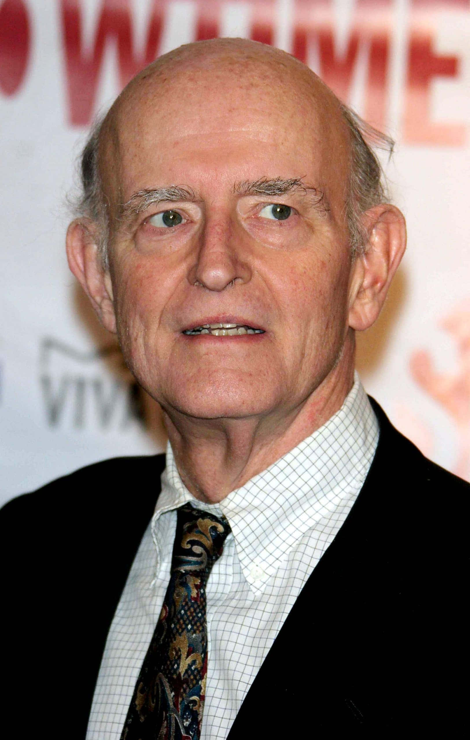 Peter Boyle 桌布 壁紙 — 一位禿頭老年男子穿著深色西裝和圖案領帶，以紅色和白色活動背景為襯的特寫肖像。