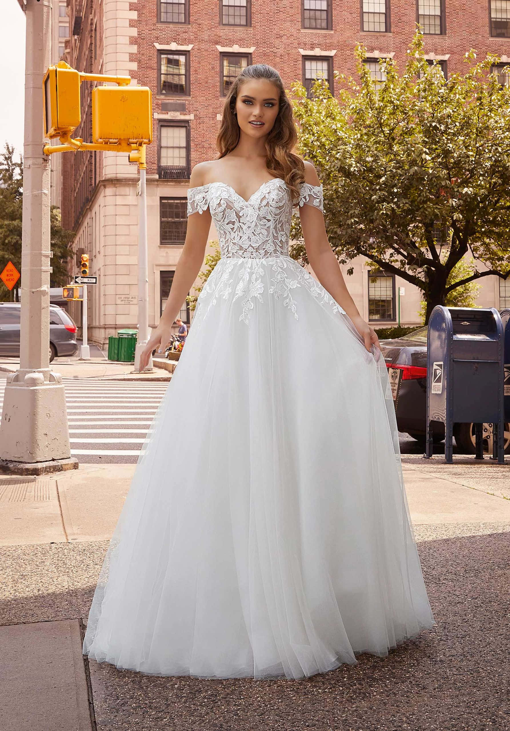 Bride's Dress 桌布 壁紙 — 一位穿著優雅白色蕾絲婚紗的女性自信地站在城市人行道上，背景是磚砌建築和綠樹。