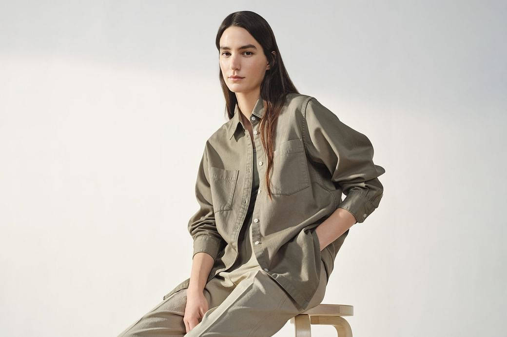 UNIQLO U by Lemaire 壁紙 — 一位穿著寬鬆橄欖色襯衫和長褲的女性，自信地坐在白色背景前的木凳上。