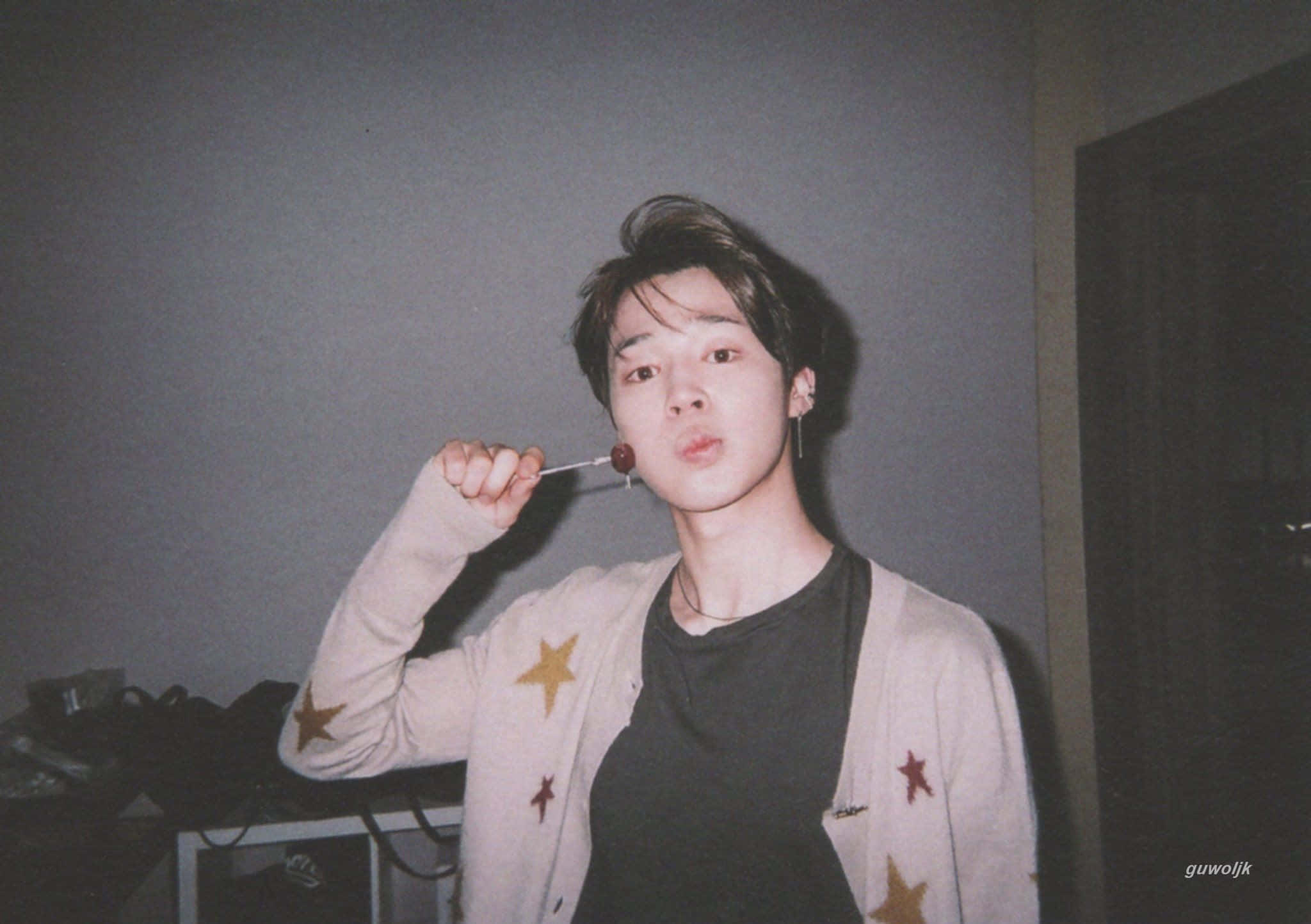 Jimin Aesthetic 桌布 壁紙 — 一位穿著星星圖案開襟毛衣的年輕男子，在室內擺姿勢，手持棒棒糖，散發柔和懷舊的光芒。