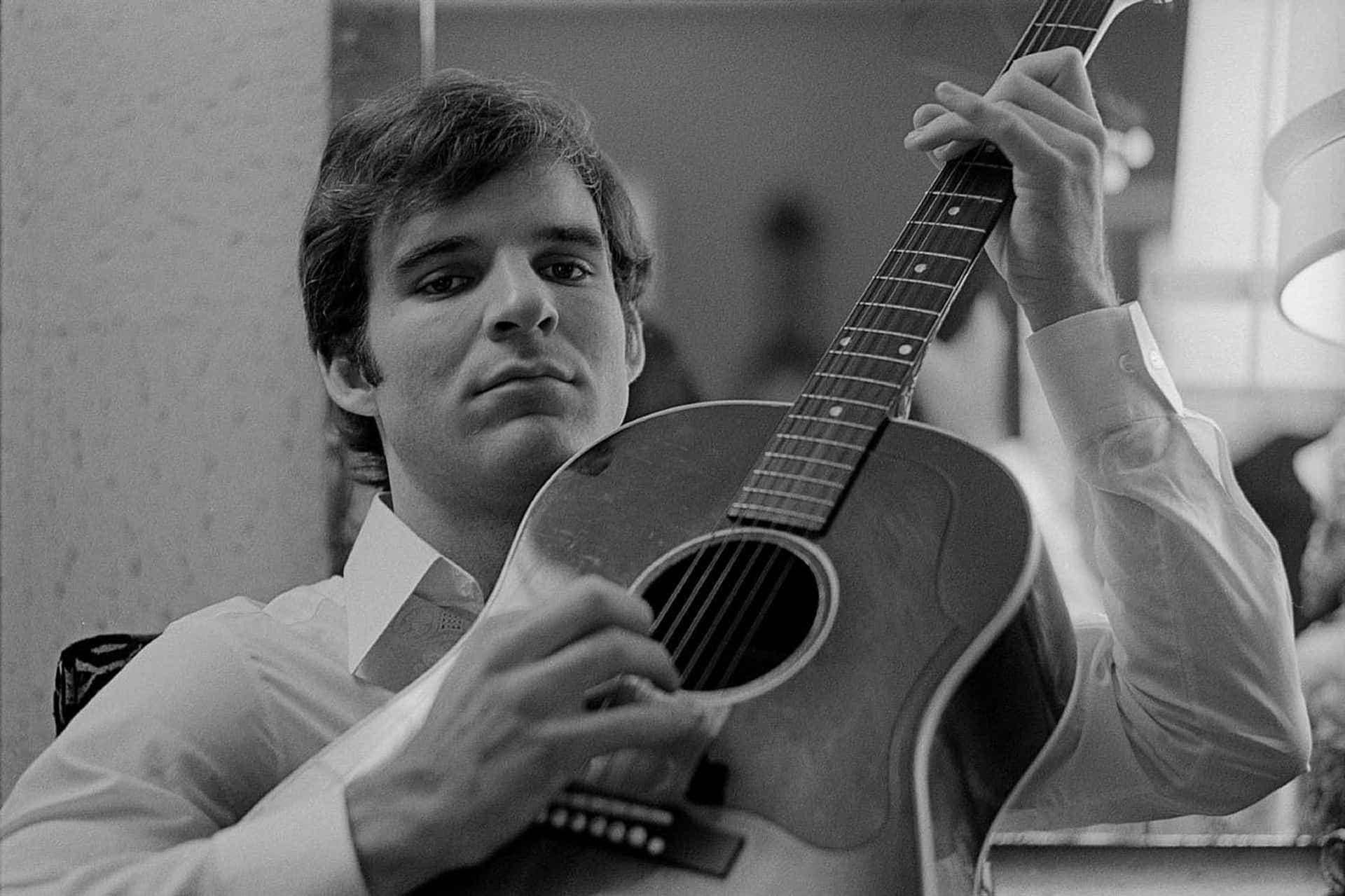 steve martin 桌布 壁紙 — 一位穿著有領襯衫的年輕男子在柔和光線的室內環境中，專注地抱著一把原聲結他。