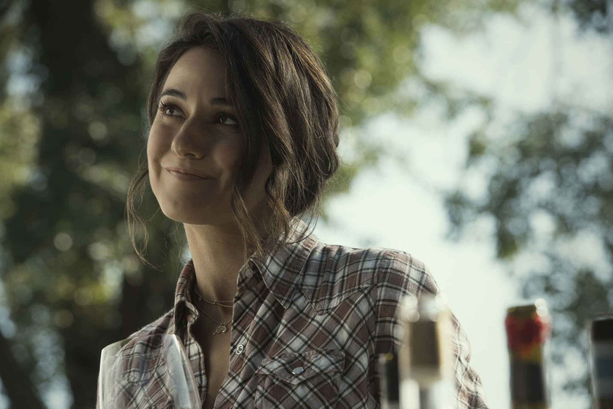 Emmanuelle Chriqui 桌布 壁紙 — 一位穿著格仔襯衫的年輕女性，在戶外樹木間灑落的斑駁陽光下溫柔微笑。