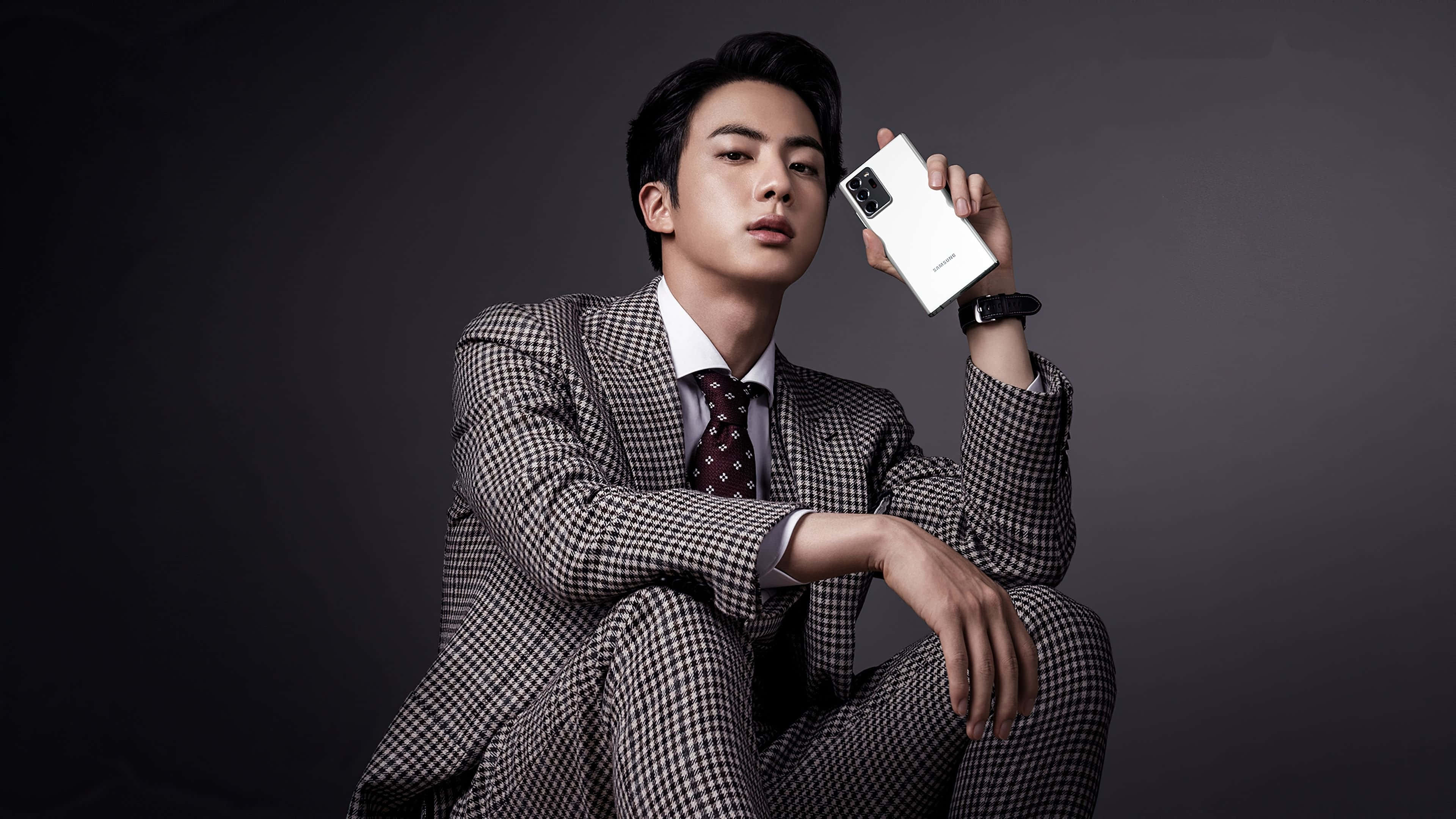 Jin 桌布 壁紙 — 一位穿著格紋西裝的時尚男士，坐在深色攝影棚背景前，手持一部白色智能手機。