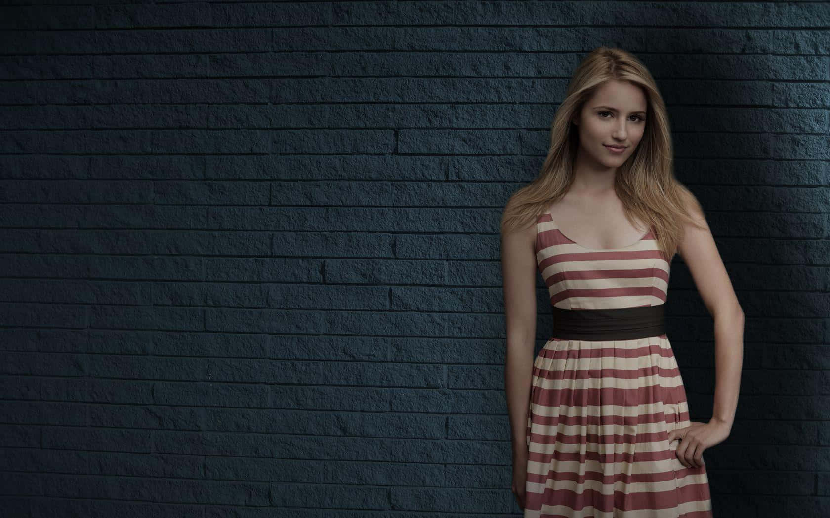dianna agron 桌布 壁紙 — 一位穿著條紋連衣裙的年輕女性在柔和的燈光下自信地站在深色紋理磚牆前。