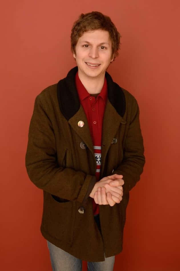 michael cera 桌布 壁紙 — 一位穿著棕色外套和紅色襯衫的年輕男子在純色陶土背景前溫暖地微笑。