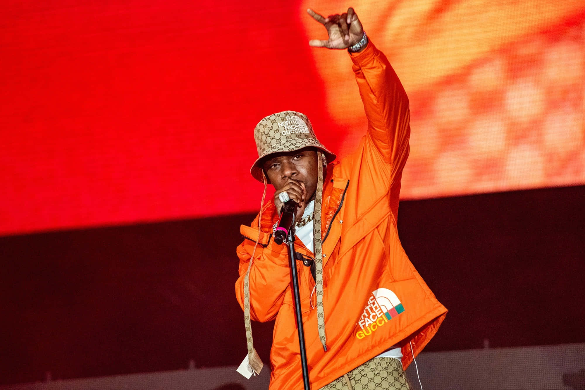 Dababy 桌布 壁紙 — 一位穿著橙色North Face外套和圖案漁夫帽的饒舌歌手在紅色和橙色LED背景的舞台上表演。