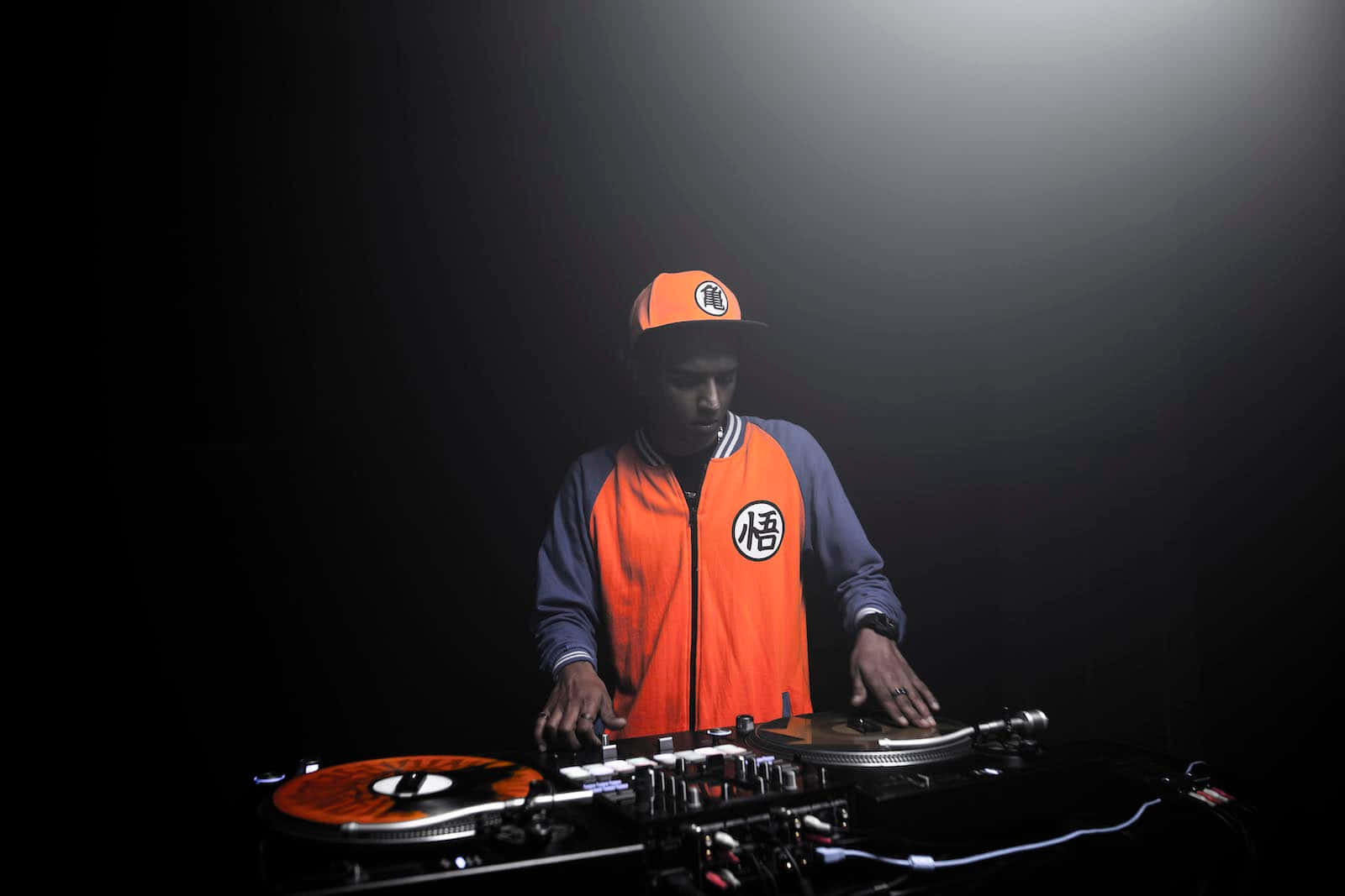 DJ 桌布 壁紙 — 一位穿著橙色悟空外套和帽子的DJ在黑暗背景的聚光燈下混音唱片。