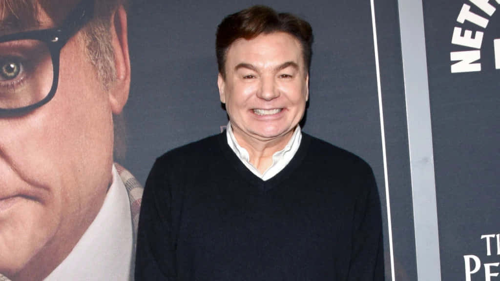 Mike Myers 桌布 壁紙 — 一位穿著深色毛衣、面帶微笑的男子站在電影海報背景前，海報上是一名戴眼鏡男子的特寫。