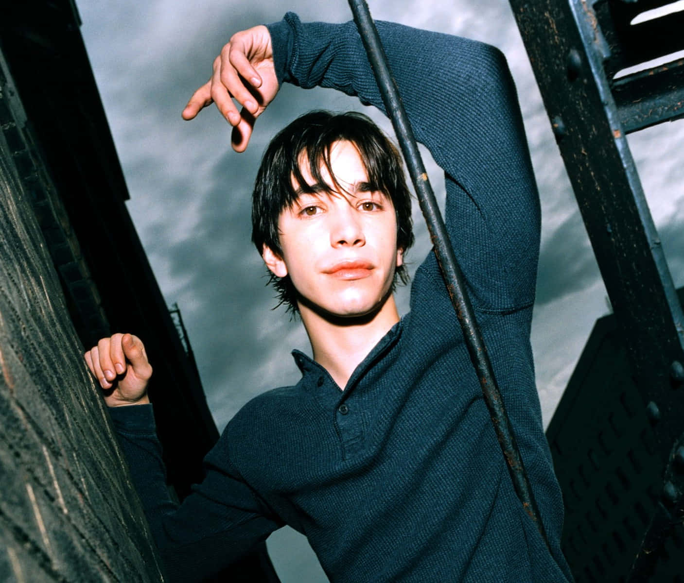 Justin Long 桌布 壁紙 — 一位穿著深色毛衣的年輕男子，在多雲的天空和戲劇性光線下，靠著逃生樓梯。