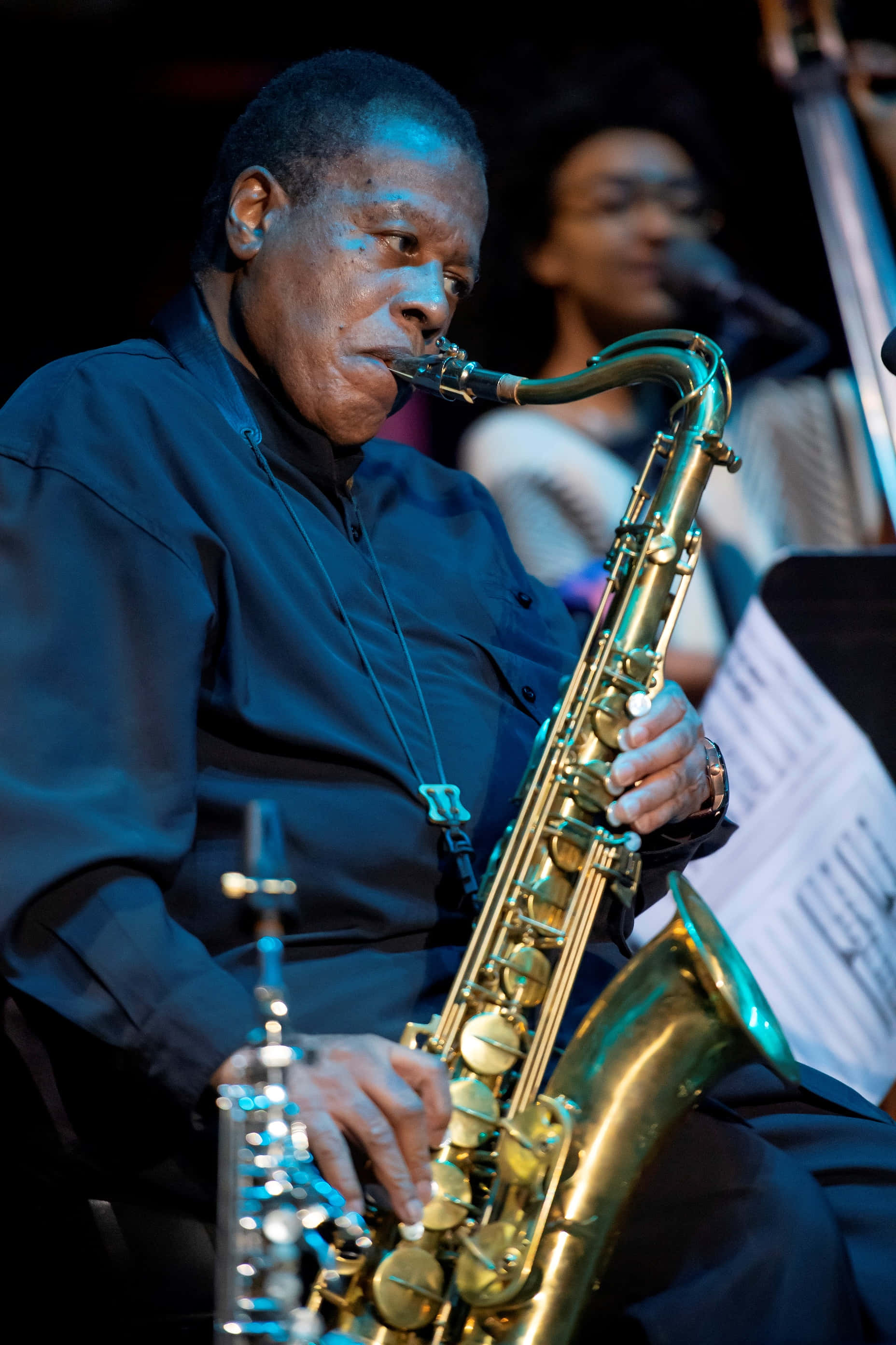 Wayne Shorter 桌布 壁紙 — 一位穿著深色襯衫的爵士薩克斯風手在舞台燈光下熱情地吹奏金色薩克斯風，身後有一位模糊的樂隊成員。