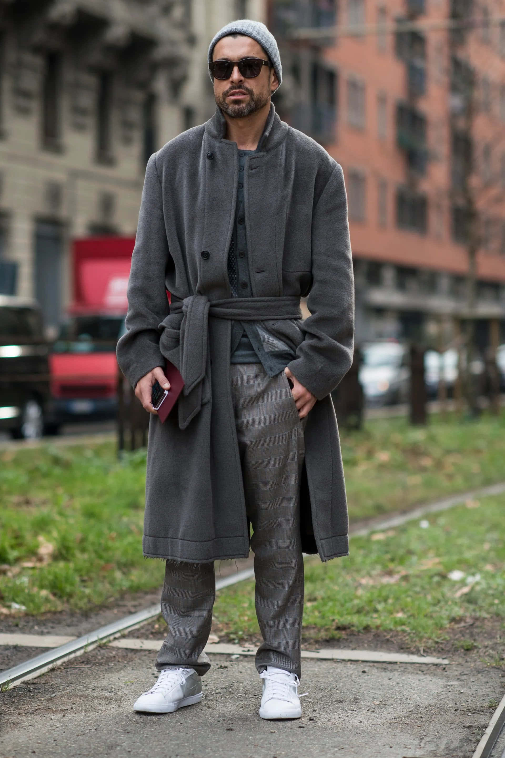 Fashion 桌布 壁紙 — 一位穿著灰色外套和冷帽的時尚男士自信地站在城市電車軌道上，背景有建築物。
