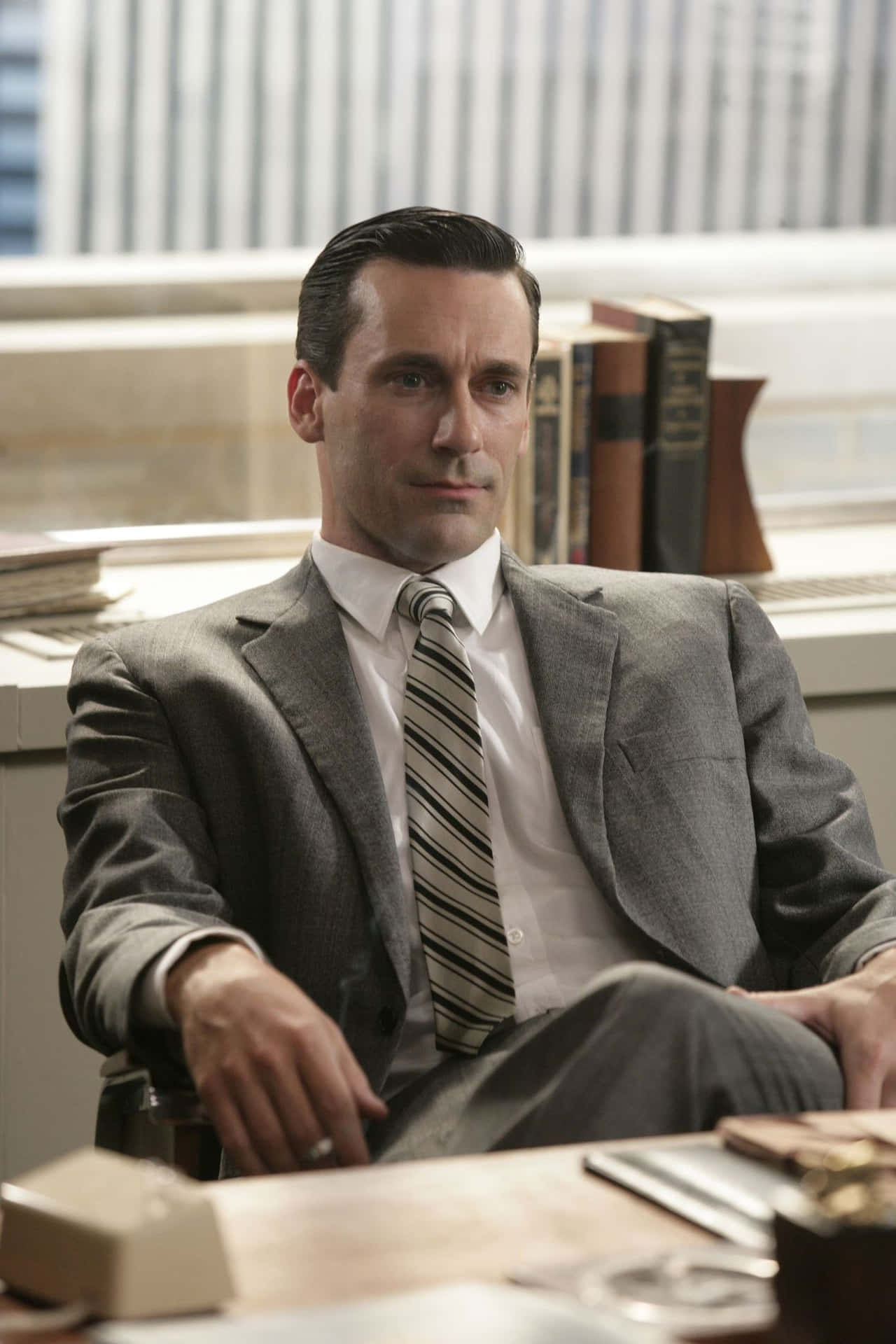 Jon Hamm 桌布 壁紙 — 一位穿著灰色西裝、衣著講究的男士坐在陽光充足的辦公室裡的木桌旁，身後可見書架和城市景觀。