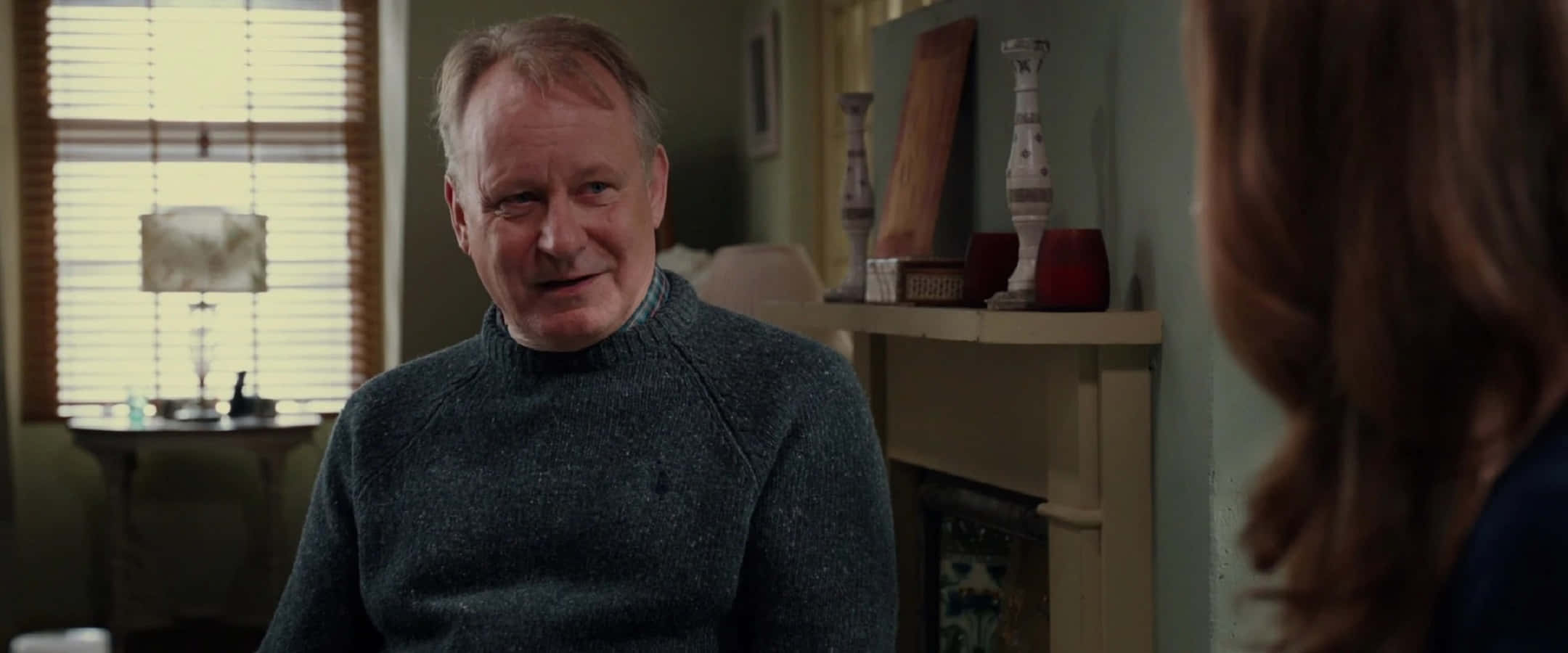 Erik Selvig 桌布 壁紙 — 一位穿著灰色高領毛衣的男子在舒適的客廳中微笑，柔和自然光透過百葉窗灑落。