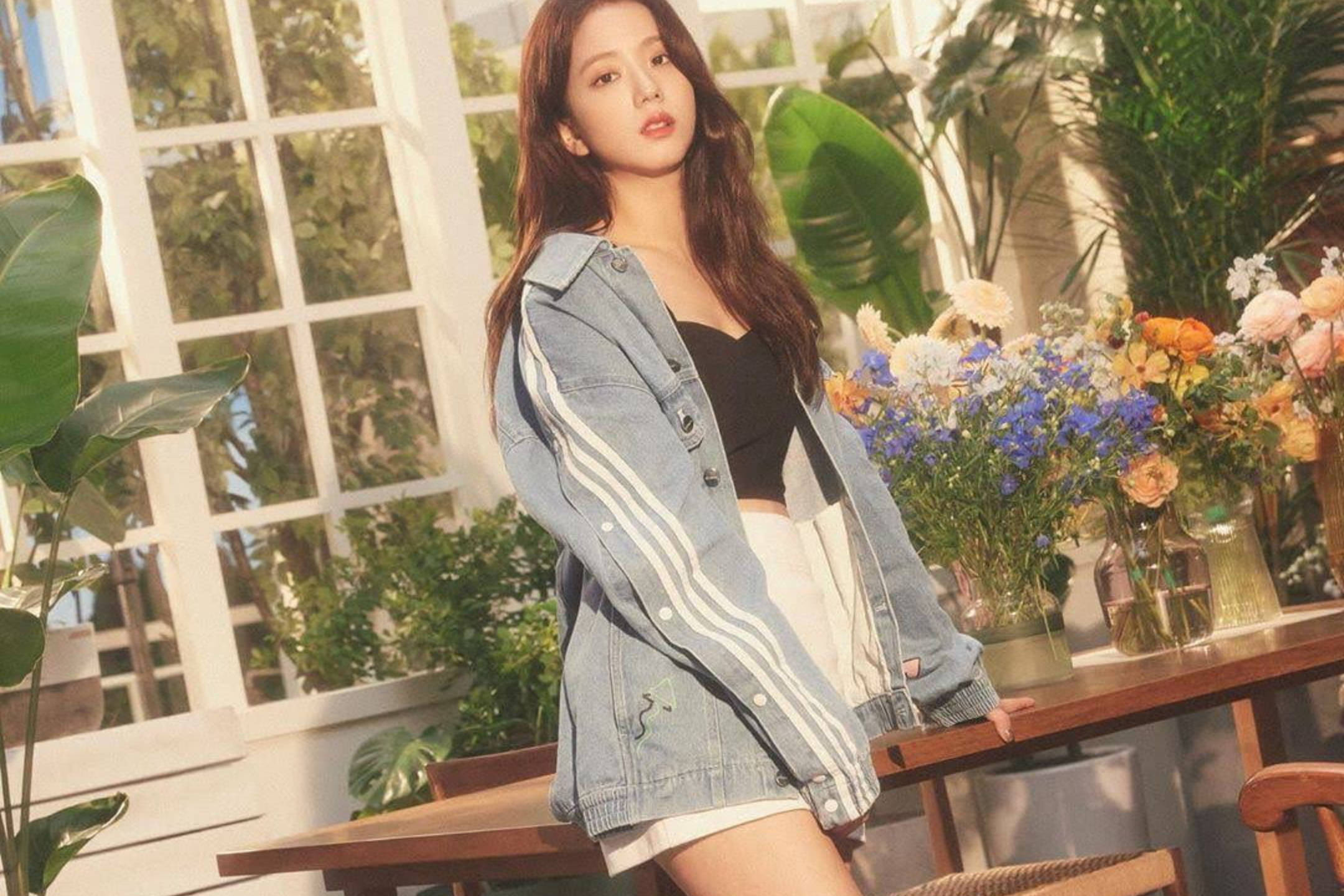 Jisoo Adidas Originals 壁紙 — 一位穿著牛仔外套的年輕女士站在窗邊，周圍環繞著茂盛的綠色植物和色彩繽紛的花藝佈置。