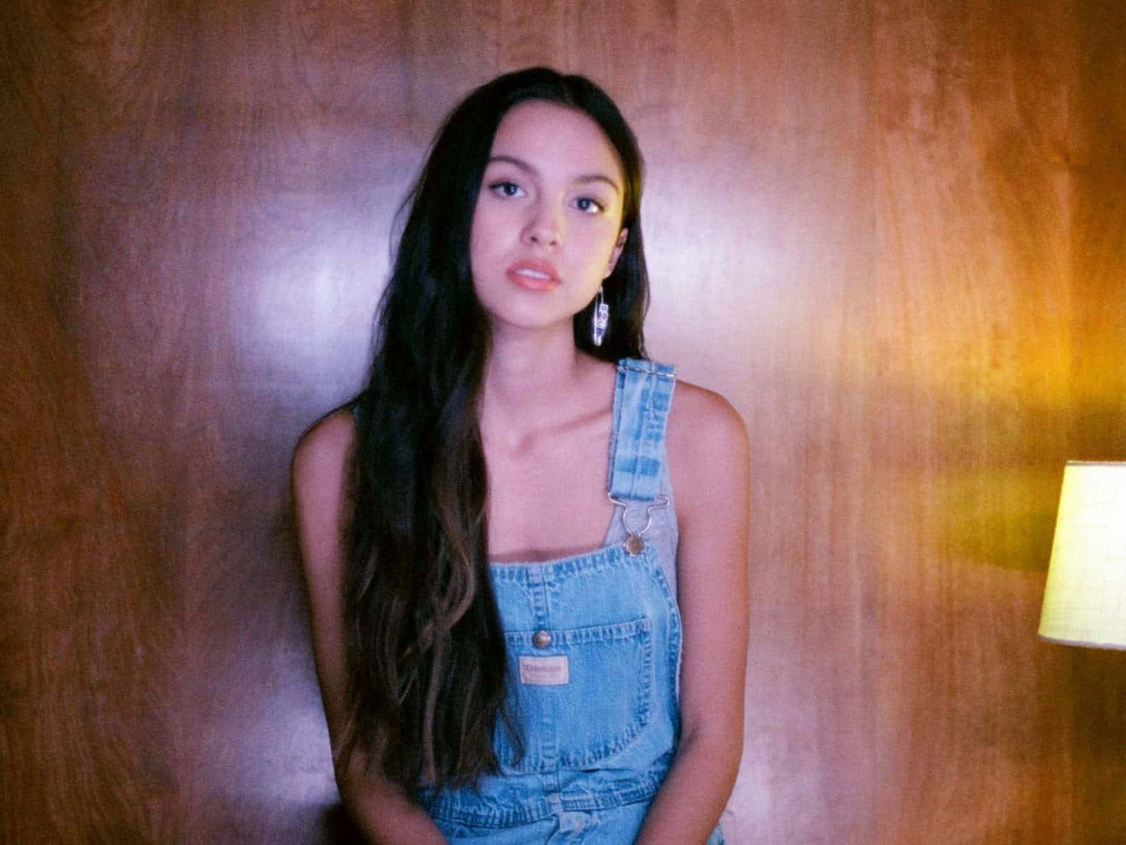 Olivia Rodrigo 桌布 壁紙 — 一位穿著牛仔工裝褲的年輕女性靠著溫暖的木牆站立，旁邊有一盞柔和的燈在發光。