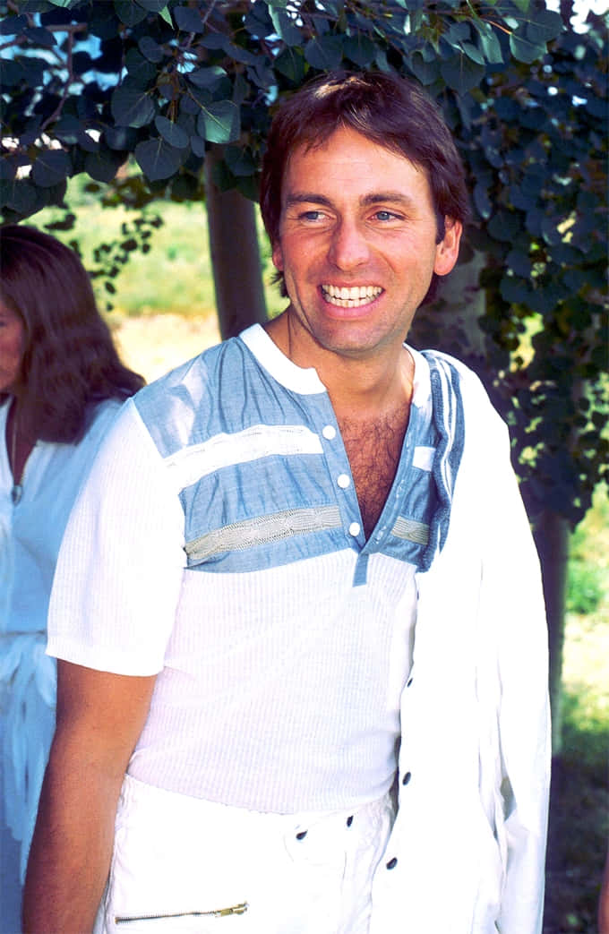 John Ritter 桌布 壁紙 — 一位穿著白藍間條襯衫的開朗男子，在陽光明媚的日子裡，在綠樹成蔭下燦爛地微笑。