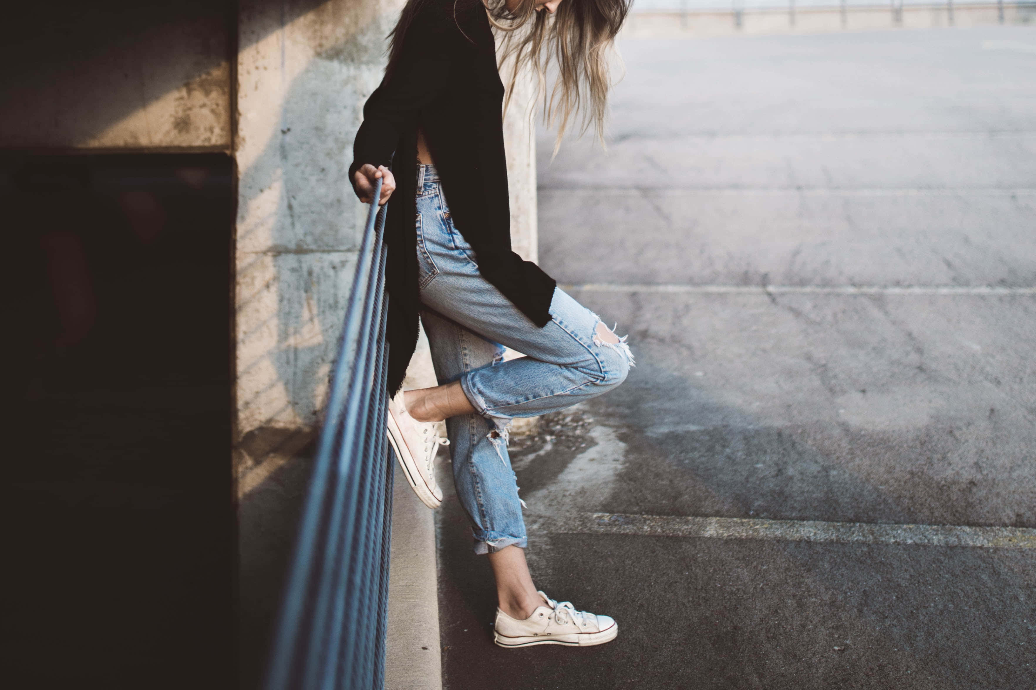 style 桌布 壁紙 — 一位穿著破洞牛仔褲和白色運動鞋的年輕女性，在沐浴著溫暖金色光線的城市環境中，隨意地靠在欄杆上。